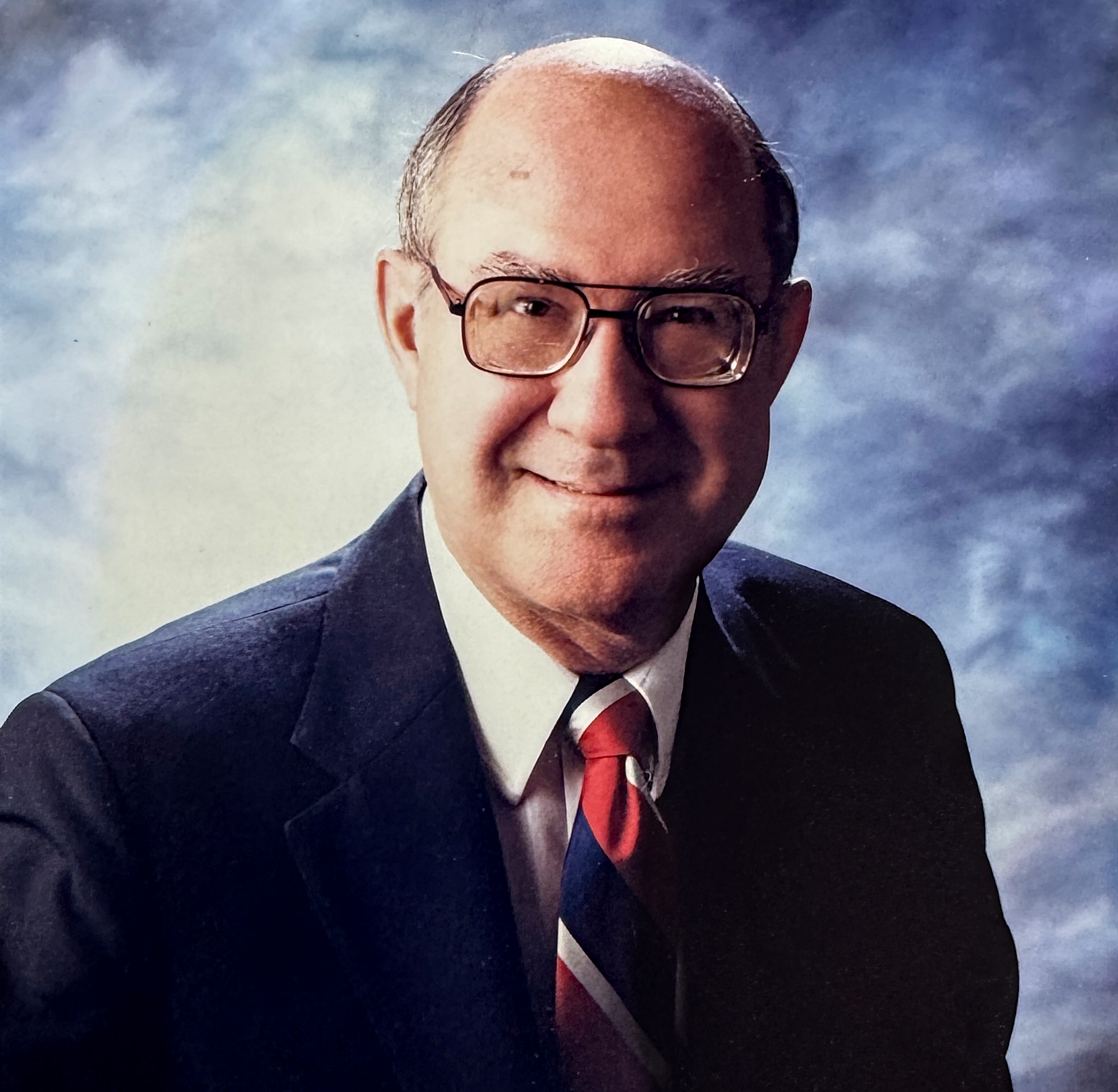Close-up of a smiling man wearing glasses, a dark suit, white shirt, and red tie, against a cloudy sky background.
