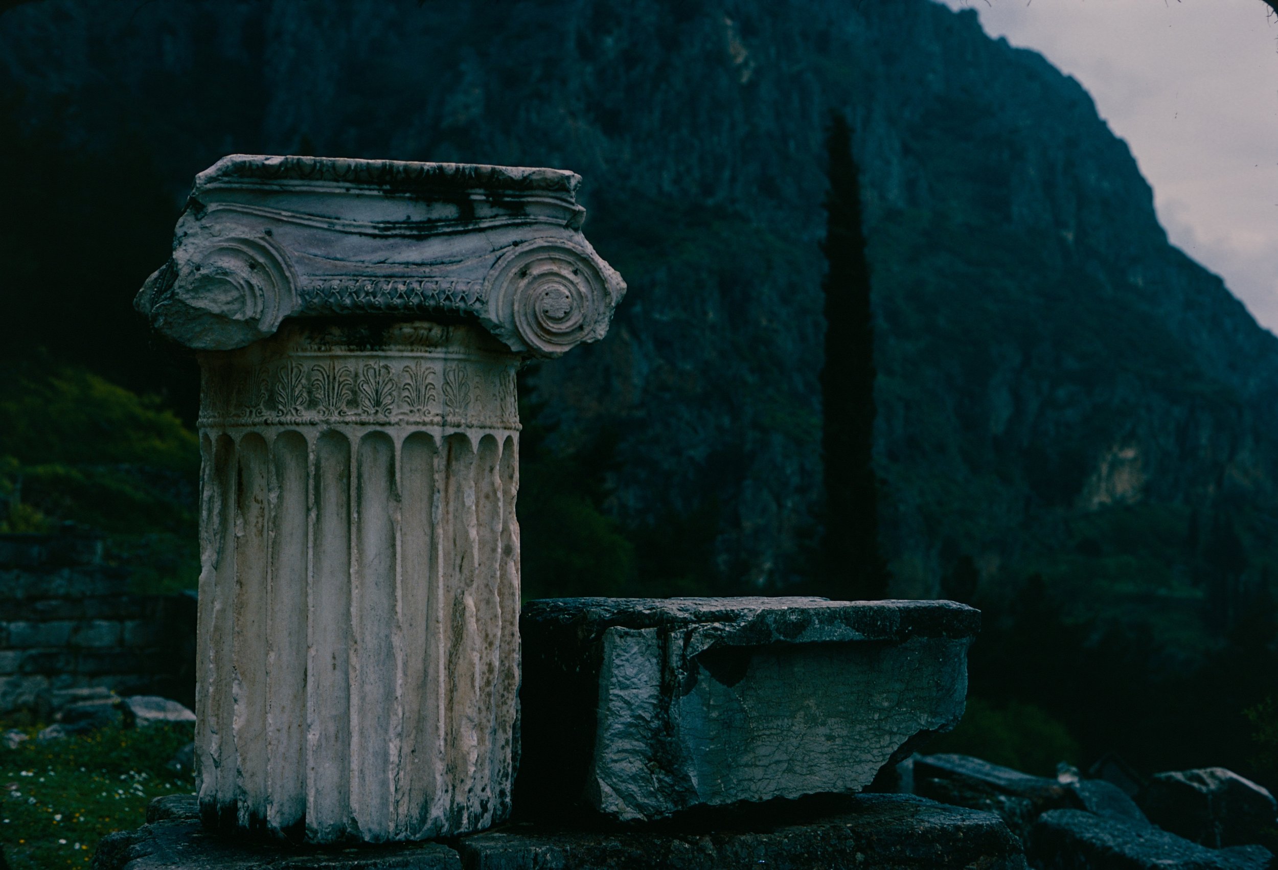 Ancient stone column with intricate carvings, set against a lush green mountainous background during dusk.