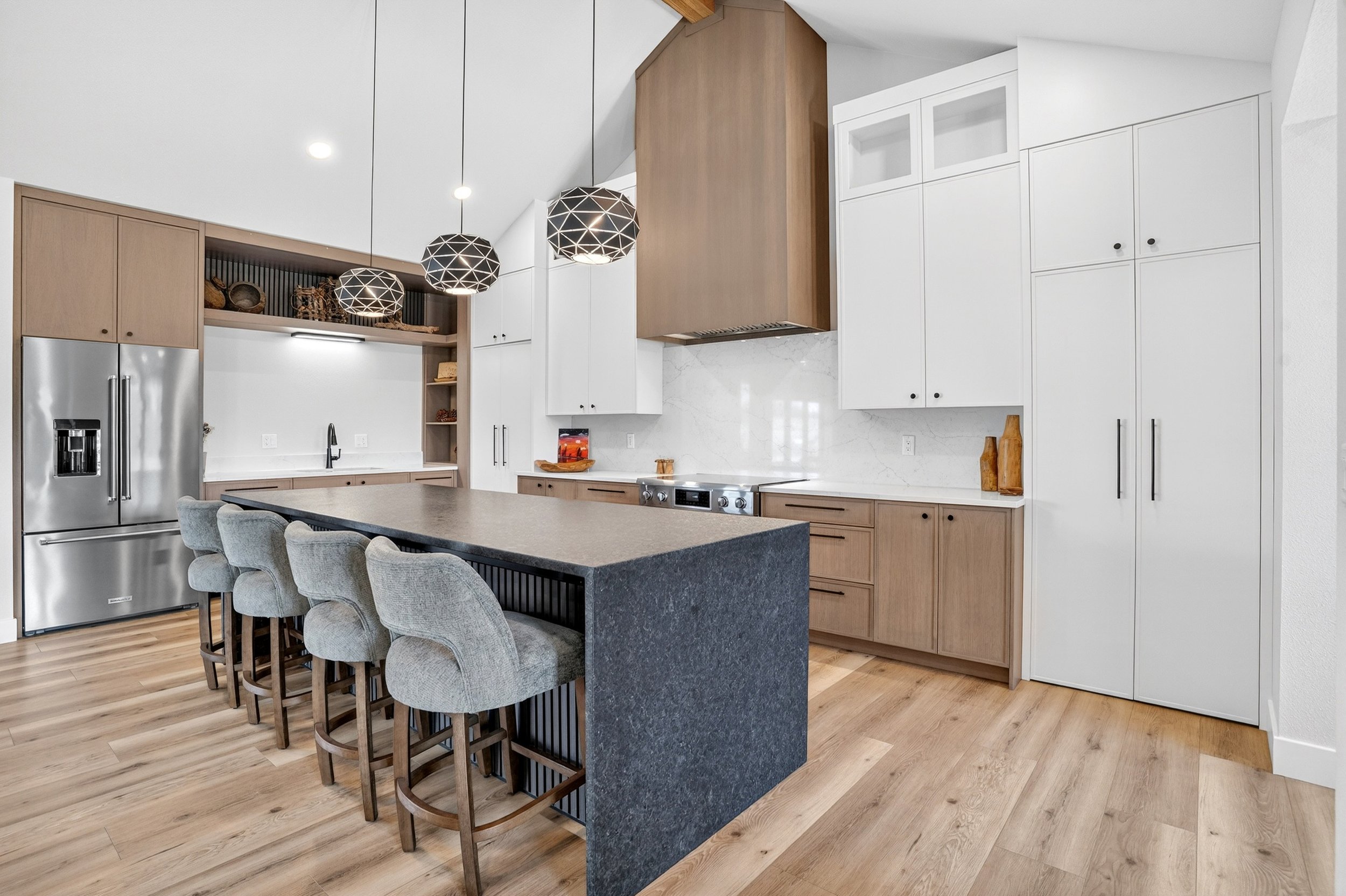 Modern kitchen with white and wood cabinetry, stainless steel refrigerator, black faucet, marble backsplash, island with gray chairs, wooden floors, and decorative hanging lights.