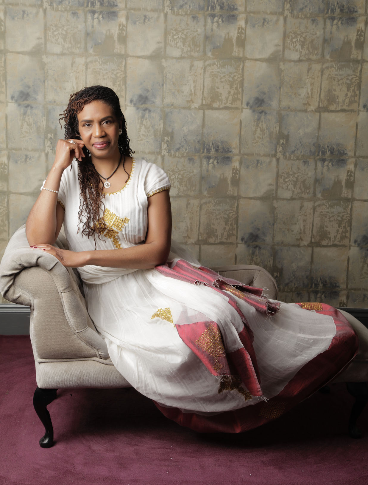 A woman with dark curly hair and brown skin sitting on a beige tufted chaise lounge. She is wearing a white dress with gold embroidery and a pink and gold patterned shawl. The woman is resting her right arm on the lounge, with her right hand touching her face, and looking at the camera. The background features a worn, textured wall in shades of beige and blue.