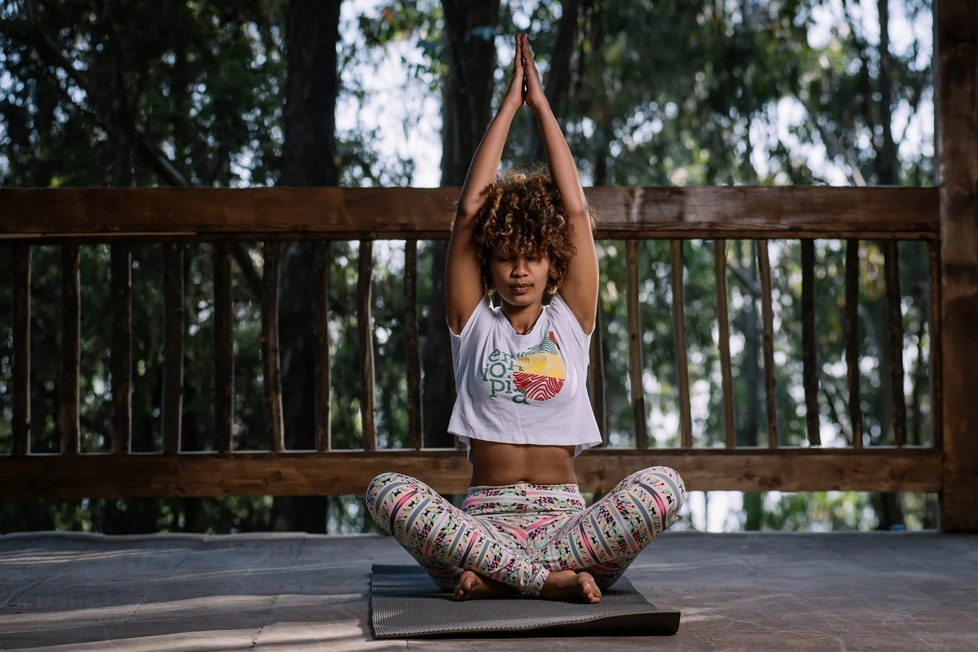 A woman practicing yoga outdoors, seated cross-legged on a yoga mat with hands raised overhead and palms together, in front of a wooden railing with trees in the background.