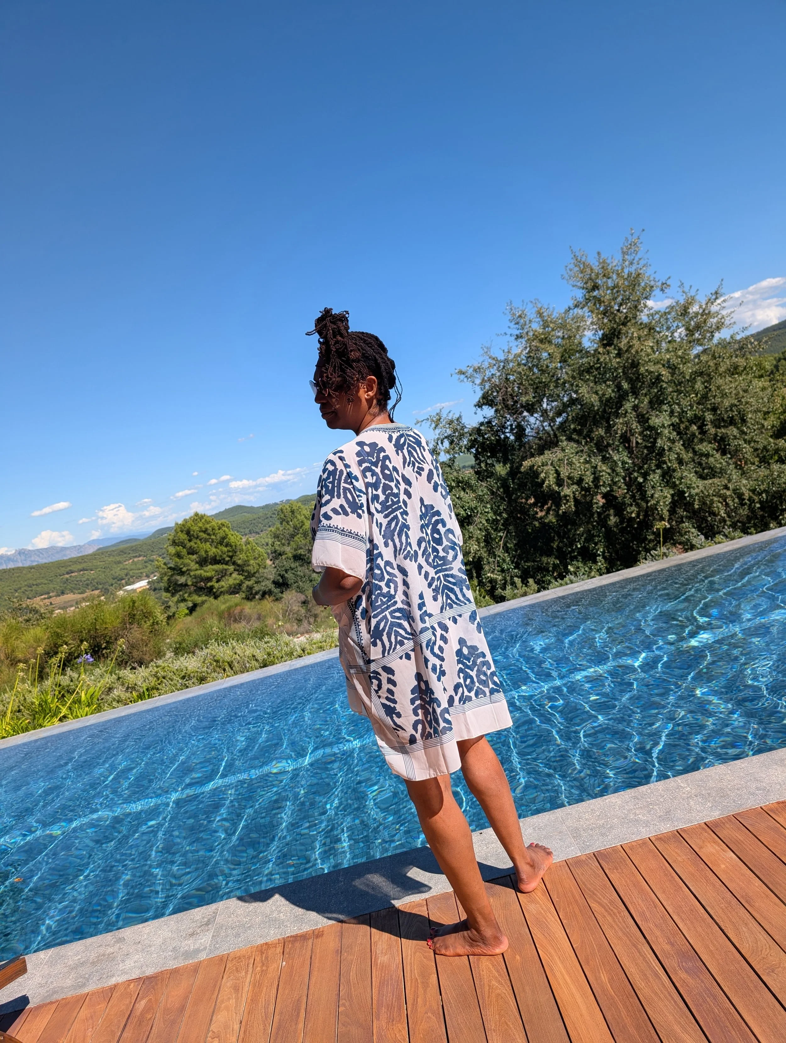 A woman with styled dreadlocks and glasses stands at the edge of a swimming pool, looking away from the camera. She is wearing a patterned cover-up. The background features green hills, trees, and a clear blue sky with some clouds.