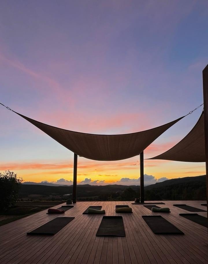 Sunset sky over an outdoor yoga deck with mats and cushions, shaded by fabric sails.