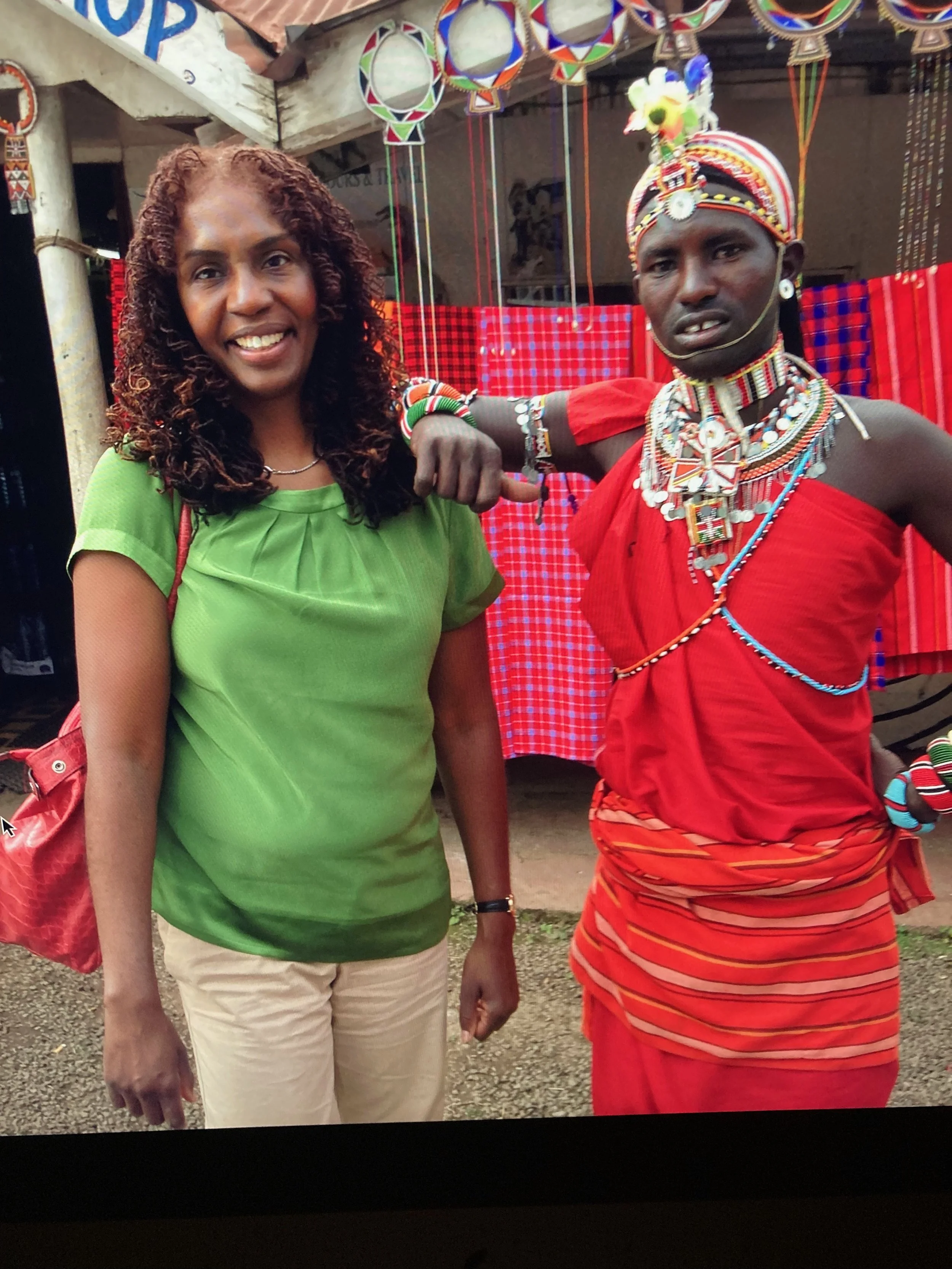 A woman smiling, wearing a green top and beige pants, standing next to a man dressed in traditional African attire with intricate jewelry and a red wrap, at an outdoor cultural event.