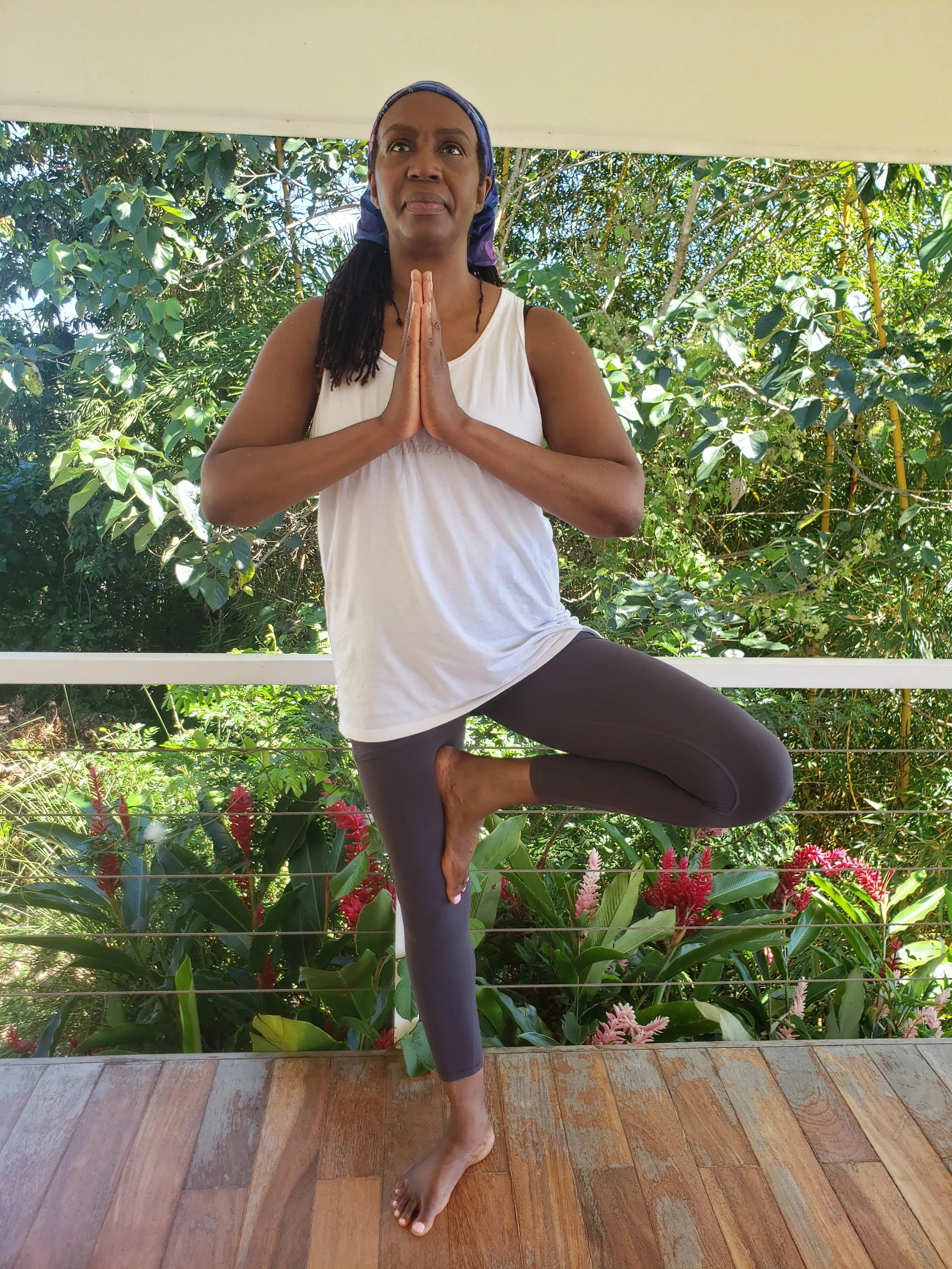 A woman practicing yoga on a wooden deck surrounded by green plants, standing in a yoga pose with hands in prayer position, one foot resting on her inner thigh.