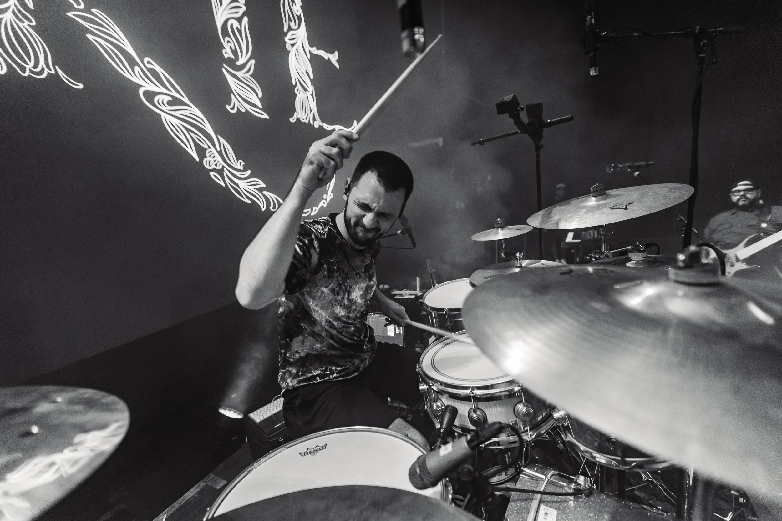 A man playing a drum set with a focused expression, holding drumsticks in a music studio or performance space, alongside a guitarist in the background, with illuminated decorative wall art visible.