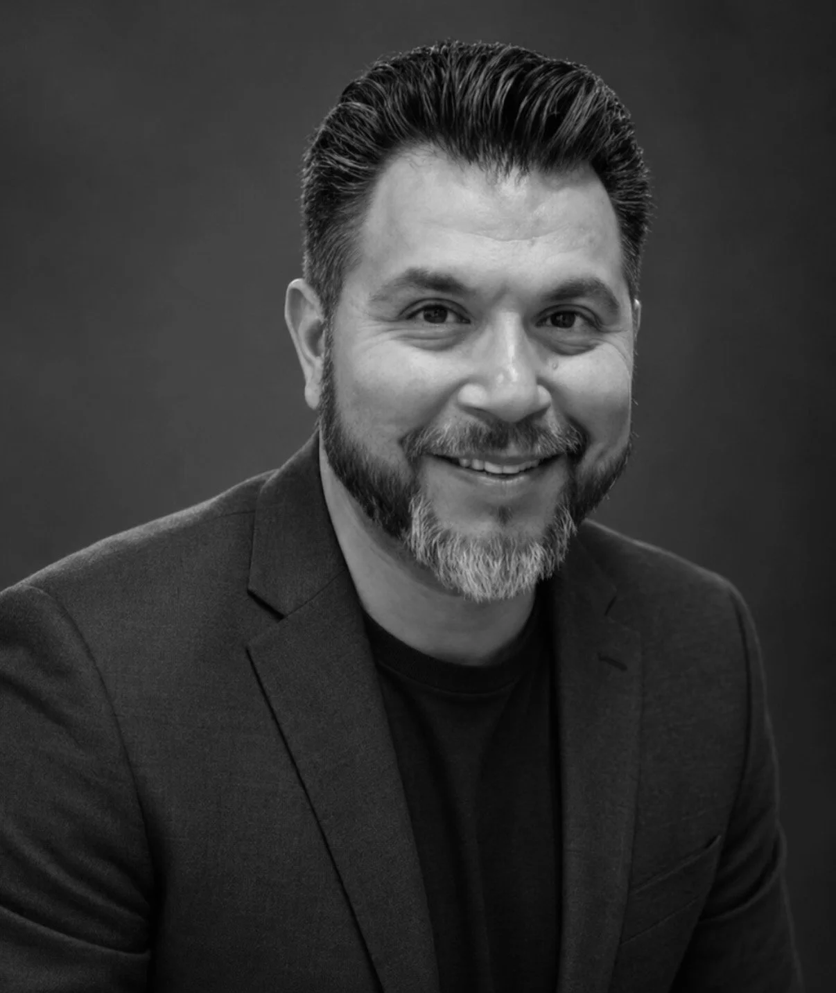 Black and white portrait of a smiling man with dark hair, beard, and mustache, wearing a suit jacket and dark shirt, against a plain dark background.