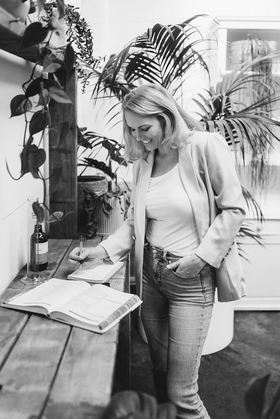 A woman with shoulder-length hair, smiling, writing in a notebook on a wooden table with an open book and a glass bottle nearby, surrounded by large potted plants.