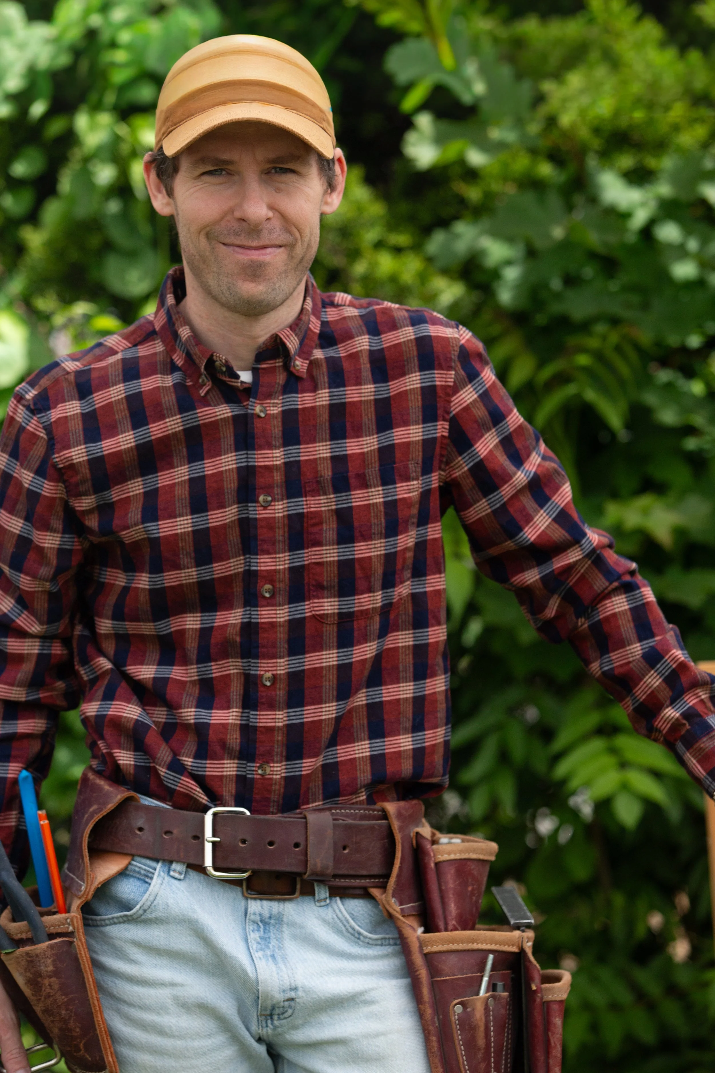 A man wearing a tan cap and a red plaid shirt standing outdoors in front of green foliage.