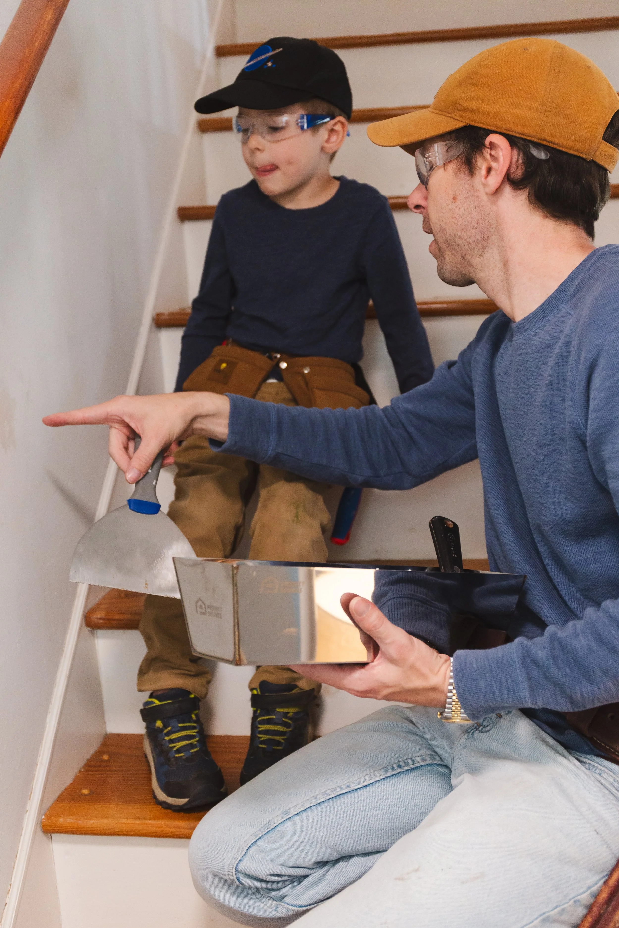 A man and a boy sitting on a staircase; the man is holding a metal container and pointing, both are wearing safety glasses and caps, and the boy is dressed in casual outdoor clothes.