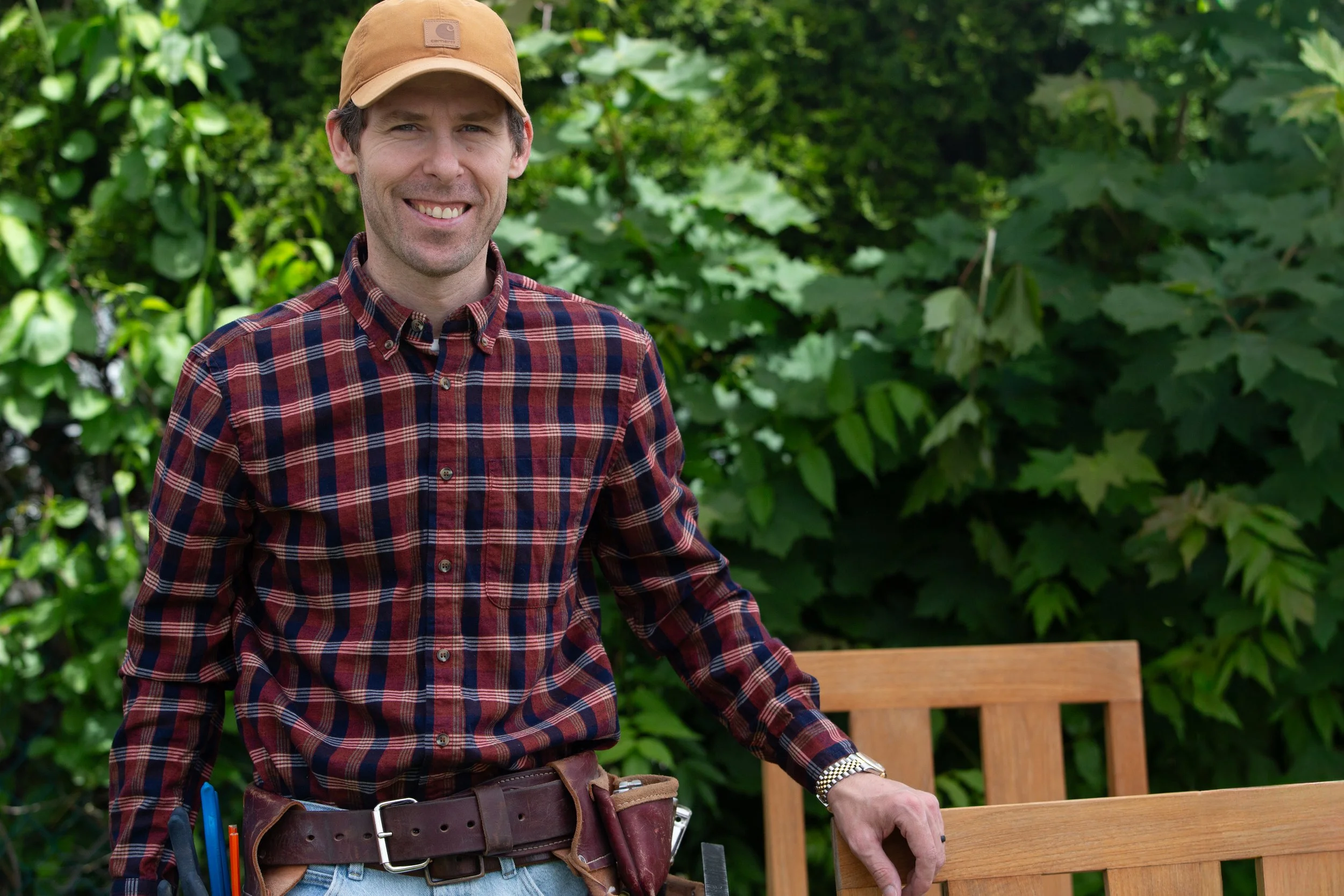 A man smiling outdoors wearing a brown cap and a red plaid shirt. He has a tool belt with tools and is standing next to a wooden bench, with lush green foliage in the background.
