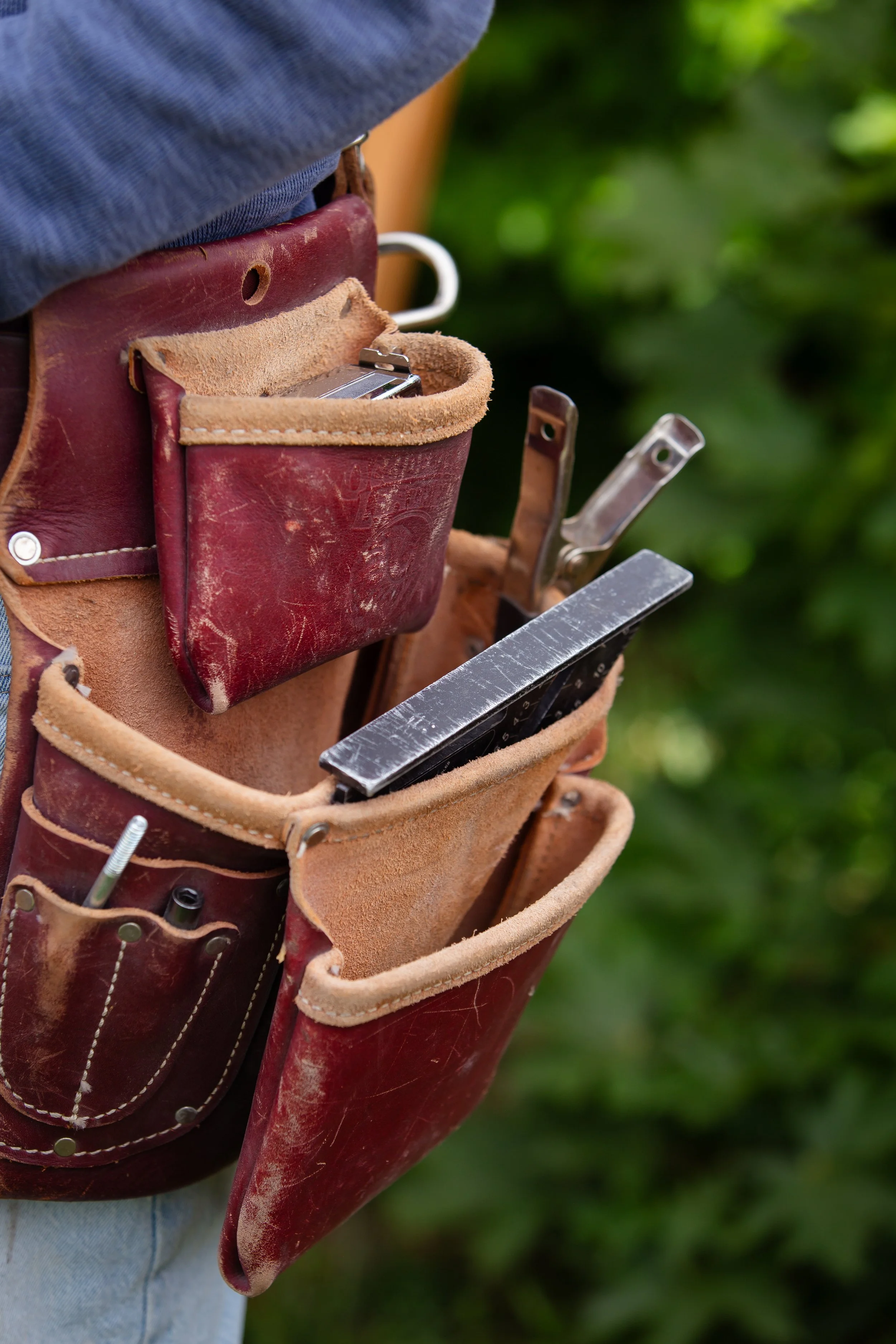 Close-up view of a person's tool belt made of leather, holding various tools, with a blurred green outdoor background.