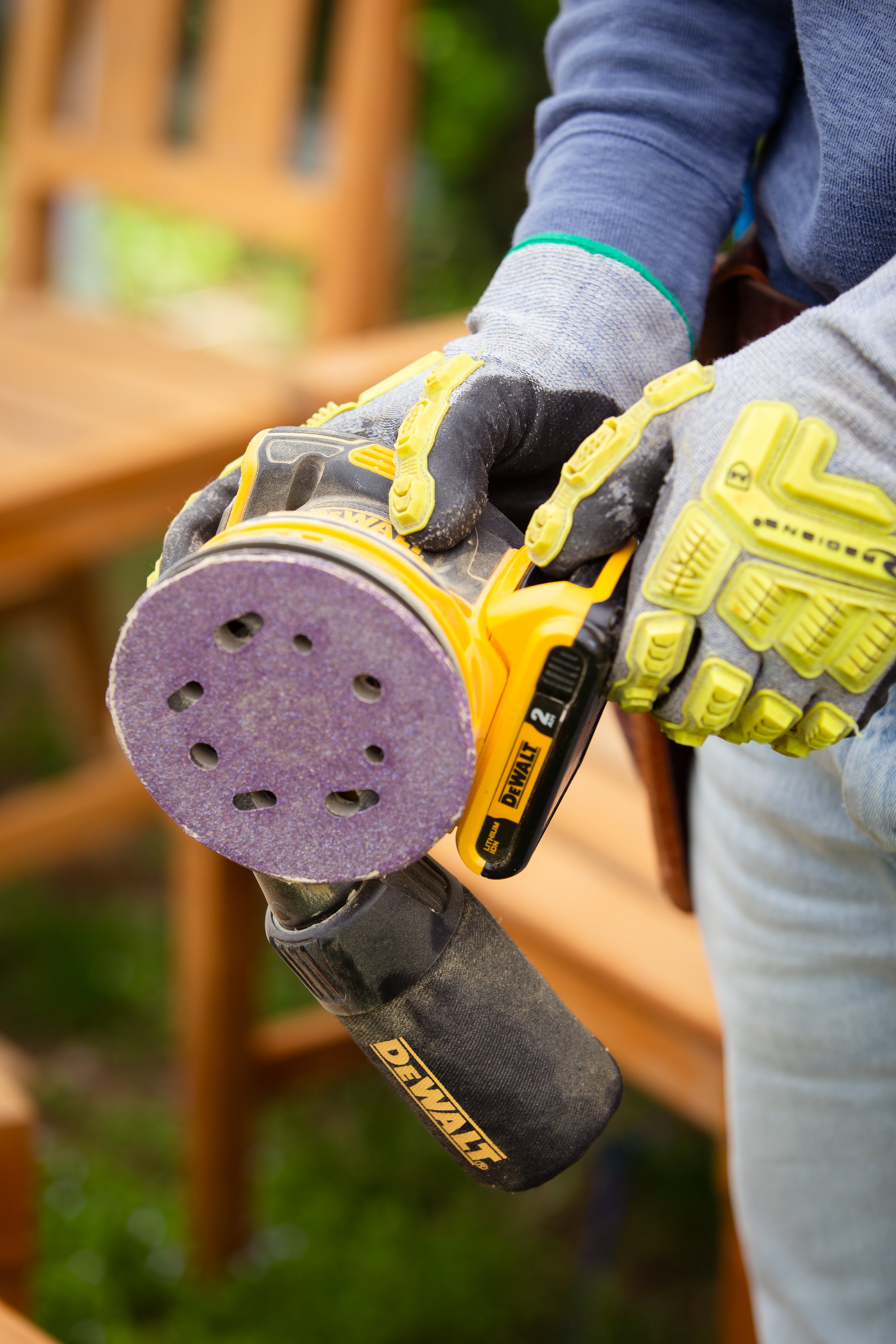 Close-up of a person holding a yellow DeWalt power sander with purple sanding pad, wearing gloves and a gray long-sleeve shirt, with a wooden structure in the background.