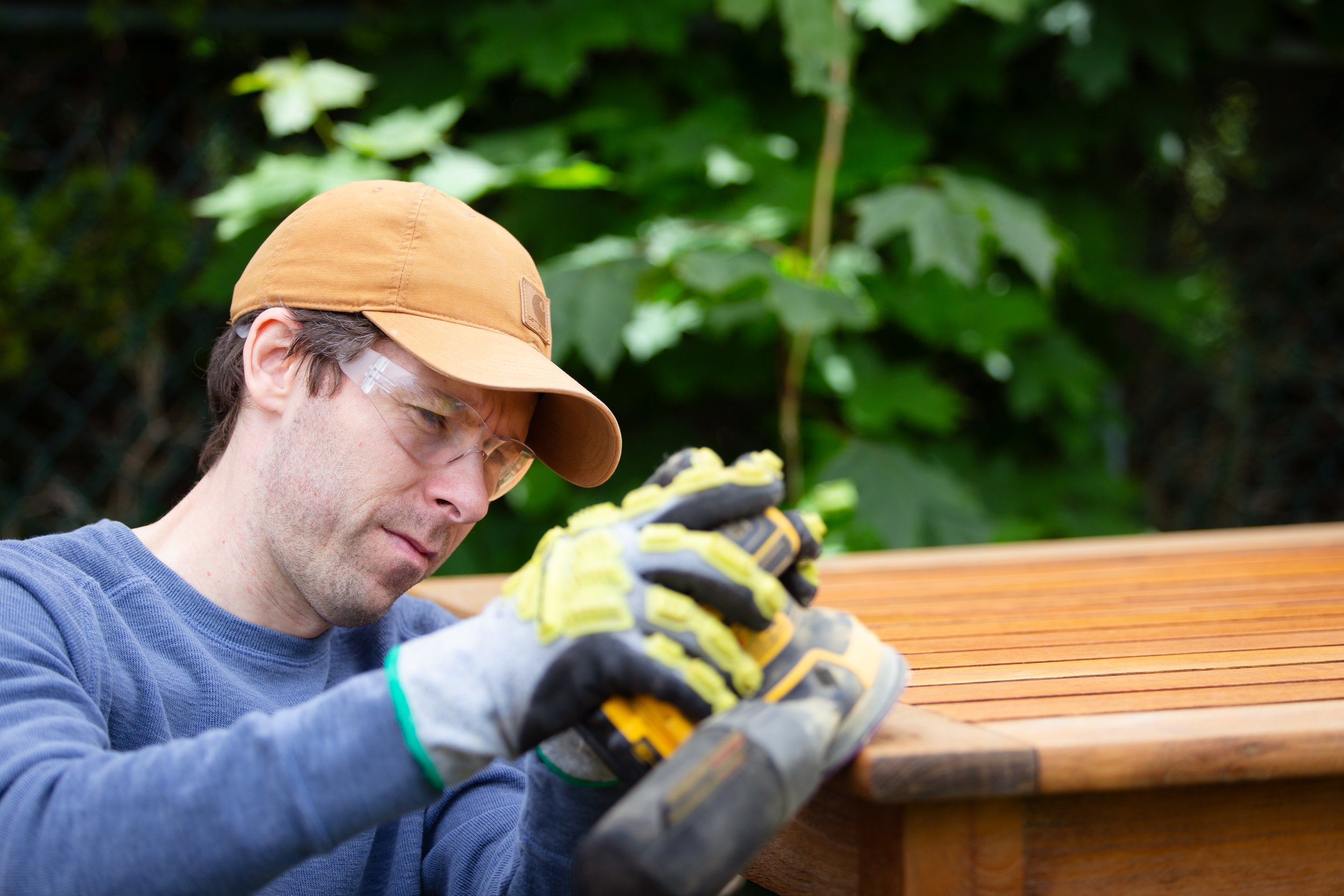 A man wearing a tan cap, safety glasses, and gloves is working on a wooden surface outdoors surrounded by green foliage.