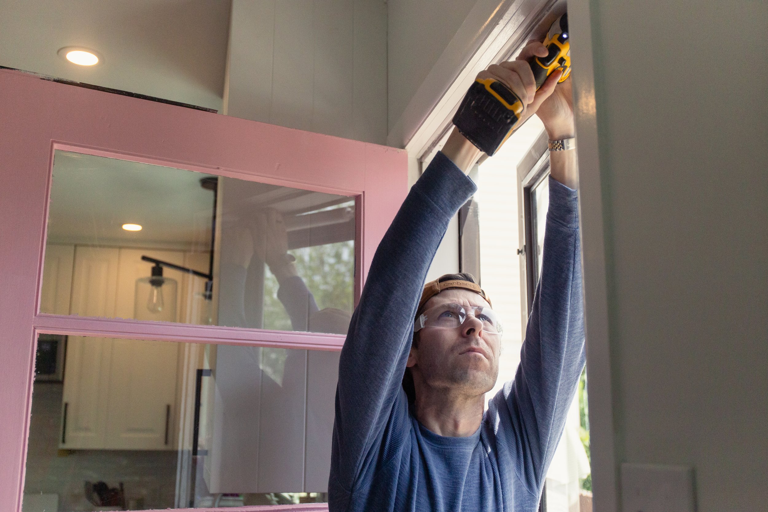 A person wearing glasses and a blue shirt is installing a window using a yellow and black power drill. They are standing indoors next to a pink-framed mirror, with the window open behind them.