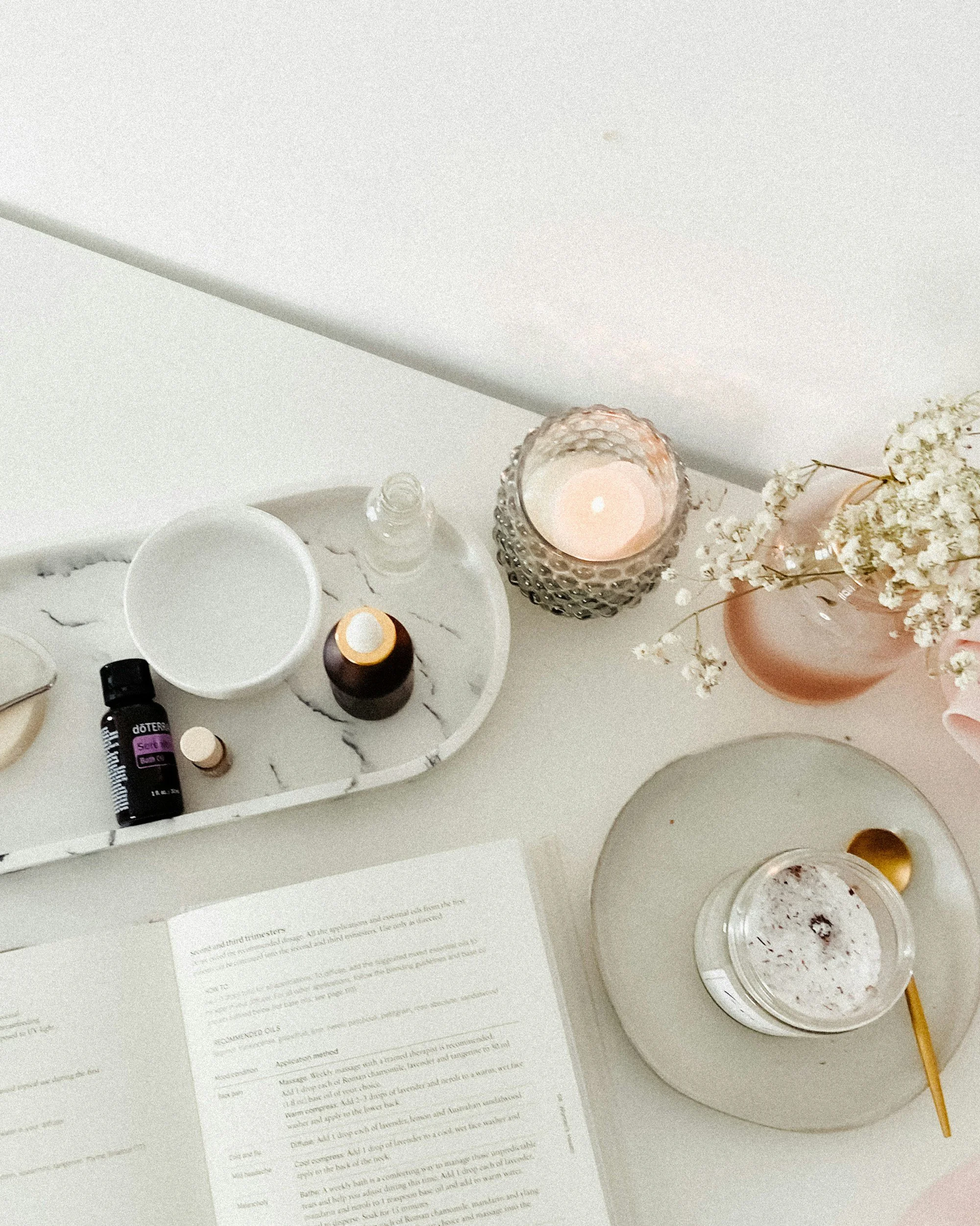 A white tray holding several small bottles, a white bowl, and a sheet of paper with printed text. Nearby, there is a lit candle in a decorative glass holder, a bouquet of white flowers in a pink vase, and part of a white round object.