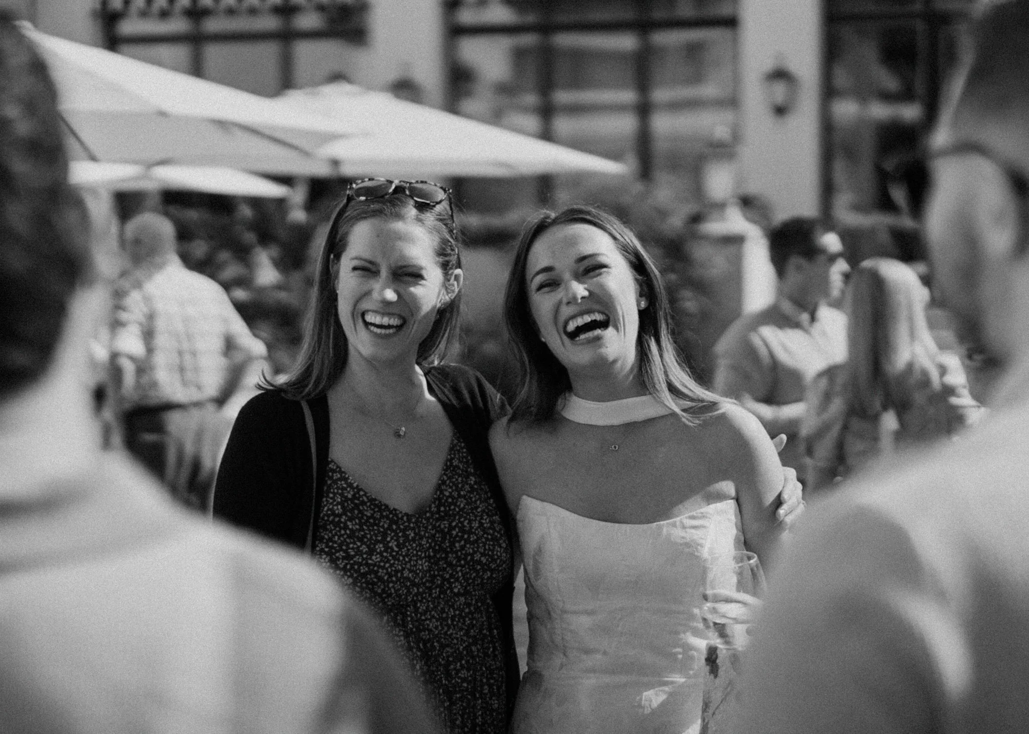 Two young women smiling and laughing at an outdoor gathering, with several people in the background and a large umbrella overhead.