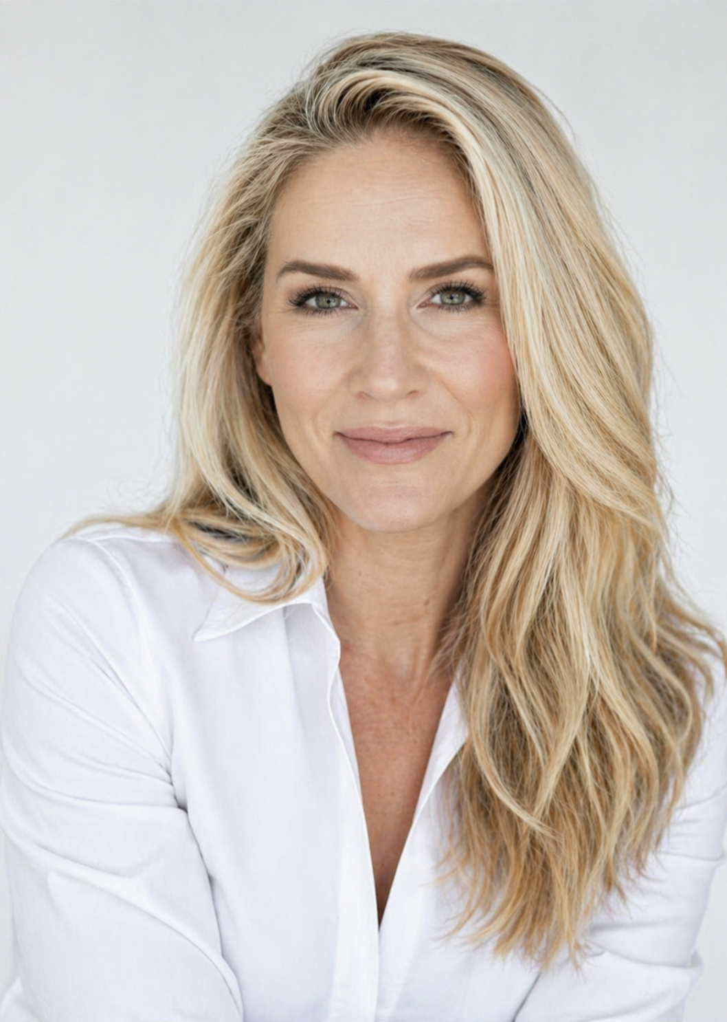 Close-up portrait of a woman with blonde wavy hair, wearing a white shirt, smiling softly against a plain light background.