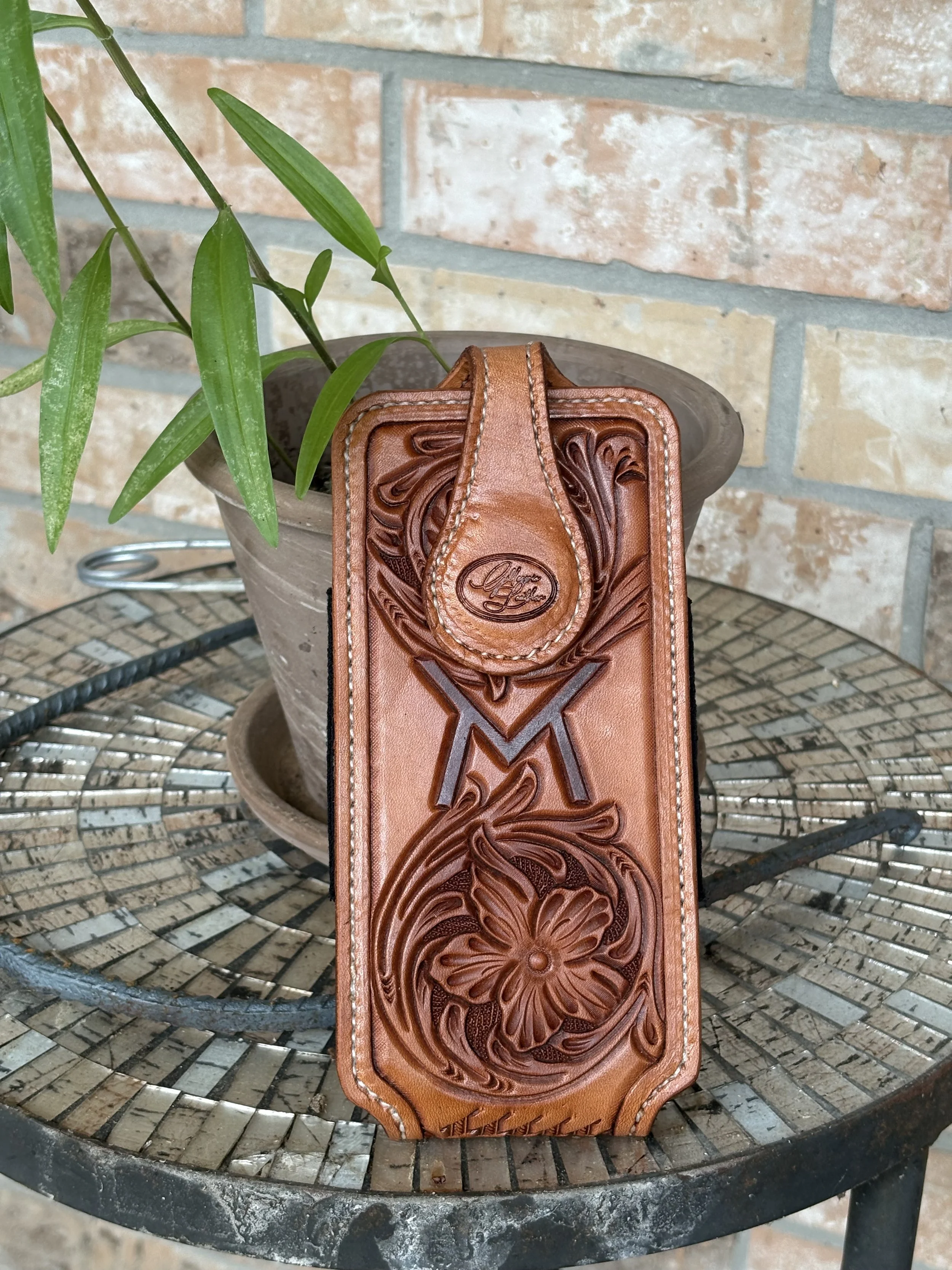 Close-up image of a brown leather wallet with intricate embossed floral and butterfly patterns, leaning against a clay flowerpot with a green plant inside, placed on a round metal table with a brick wall background.