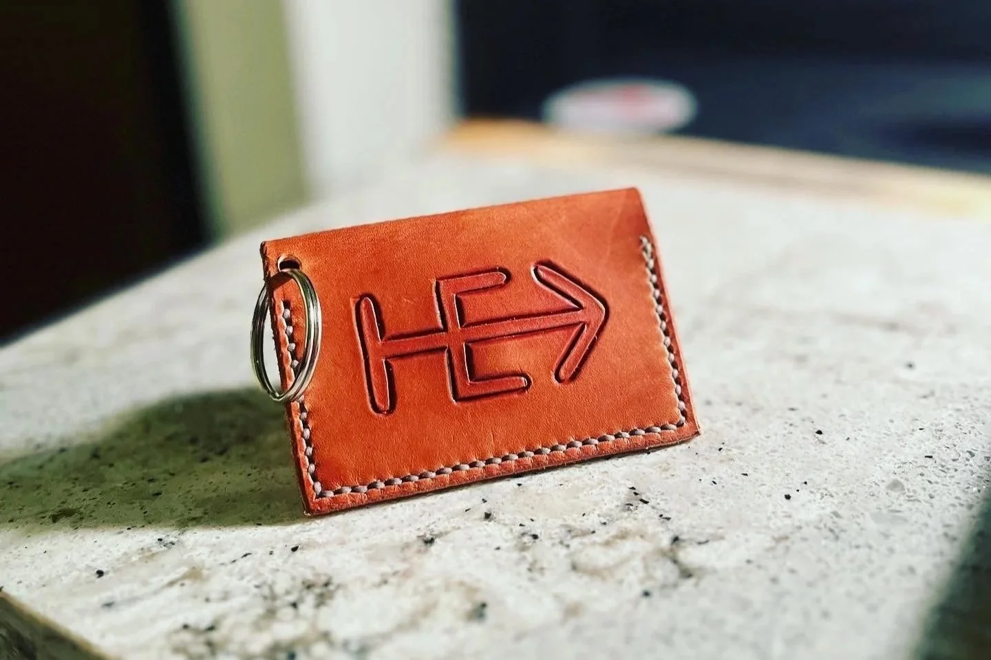 A brown leather keychain with the letters 'HE' and an arrow engraved on it, resting on a light-colored surface with a shadow cast beside it.