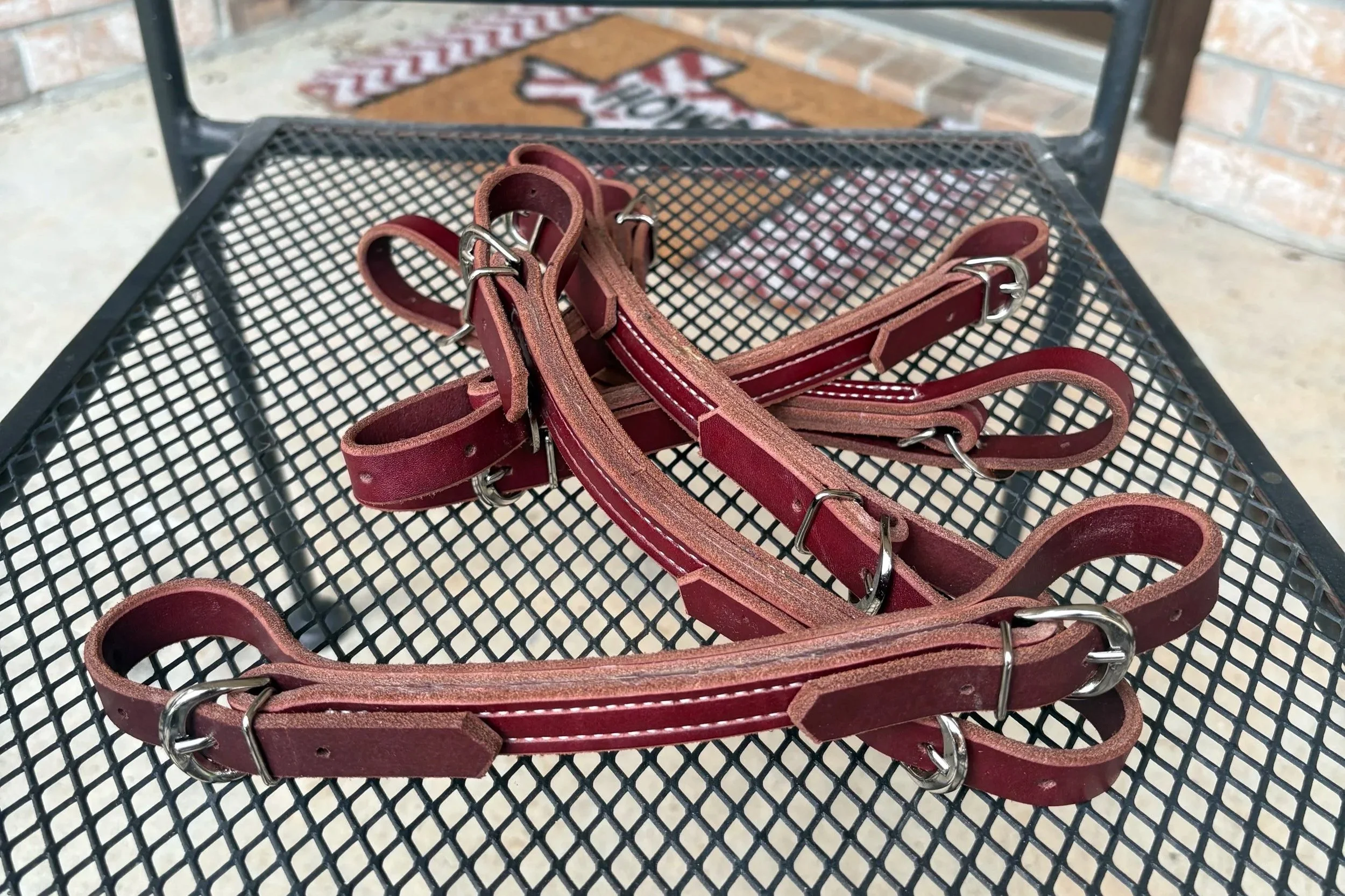 A pile of red leather dog harnesses with metal buckles on a black wire table outside, with a brick wall and a doormat in the background.