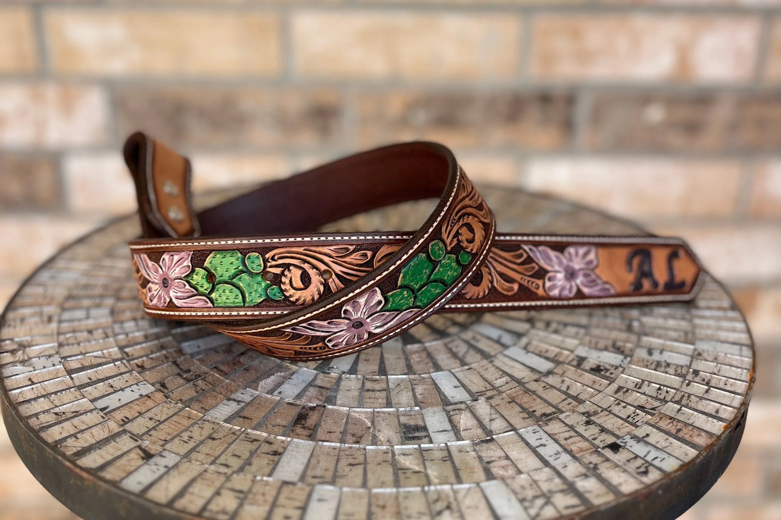 A decorative belt with intricate floral and leaf patterns in pink, purple, and green, laid on a mosaic-tiled round table with a brick wall background.