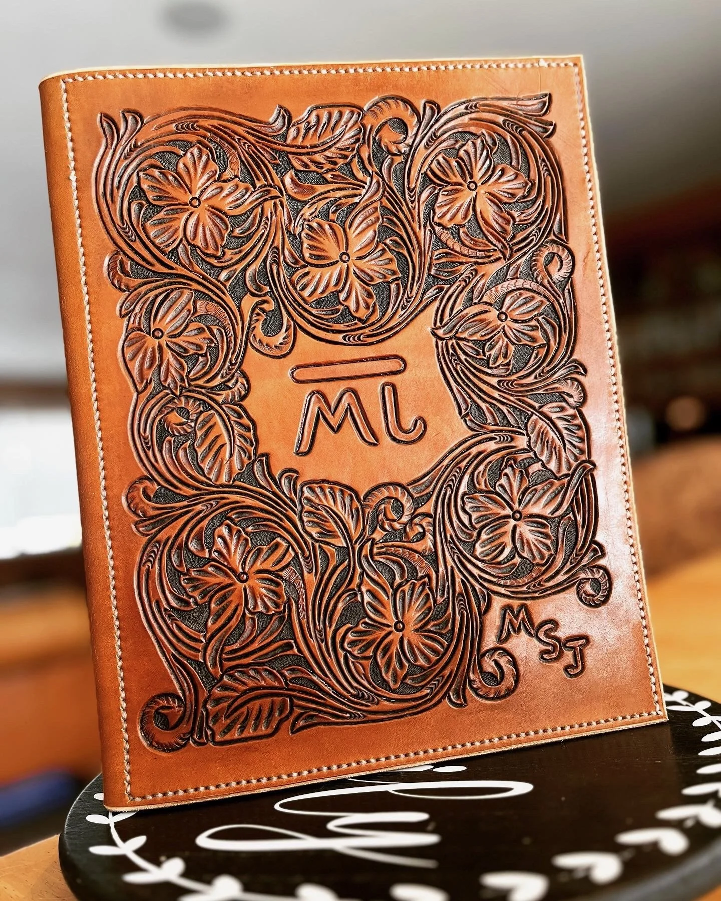 Close-up of a leather-bound notebook with intricate floral carving on the cover, featuring the initials 'ML' and 'MJ' inscribed on it.
