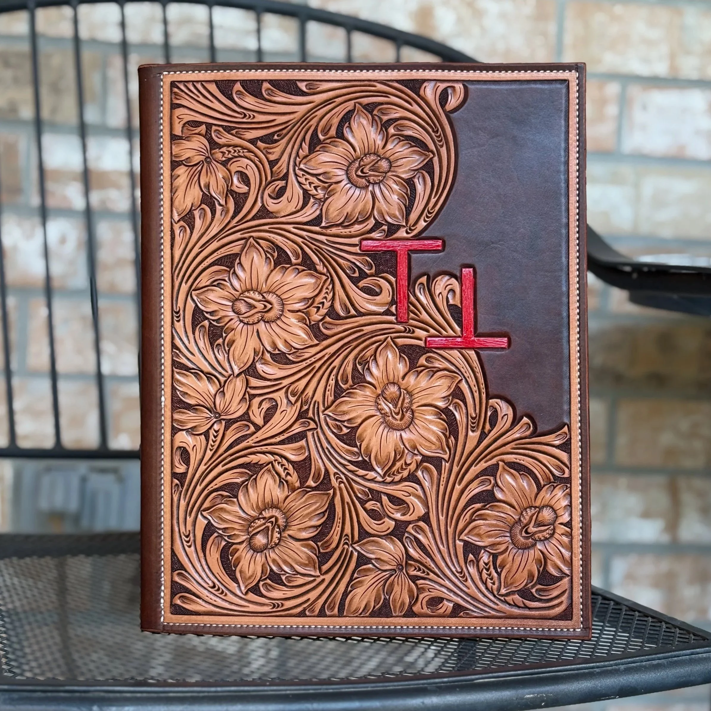 Close-up of a leather-bound book with intricate floral carving on its cover, featuring large flowers and swirling leaves, situated on a metal outdoor table with a brick wall in the background.
