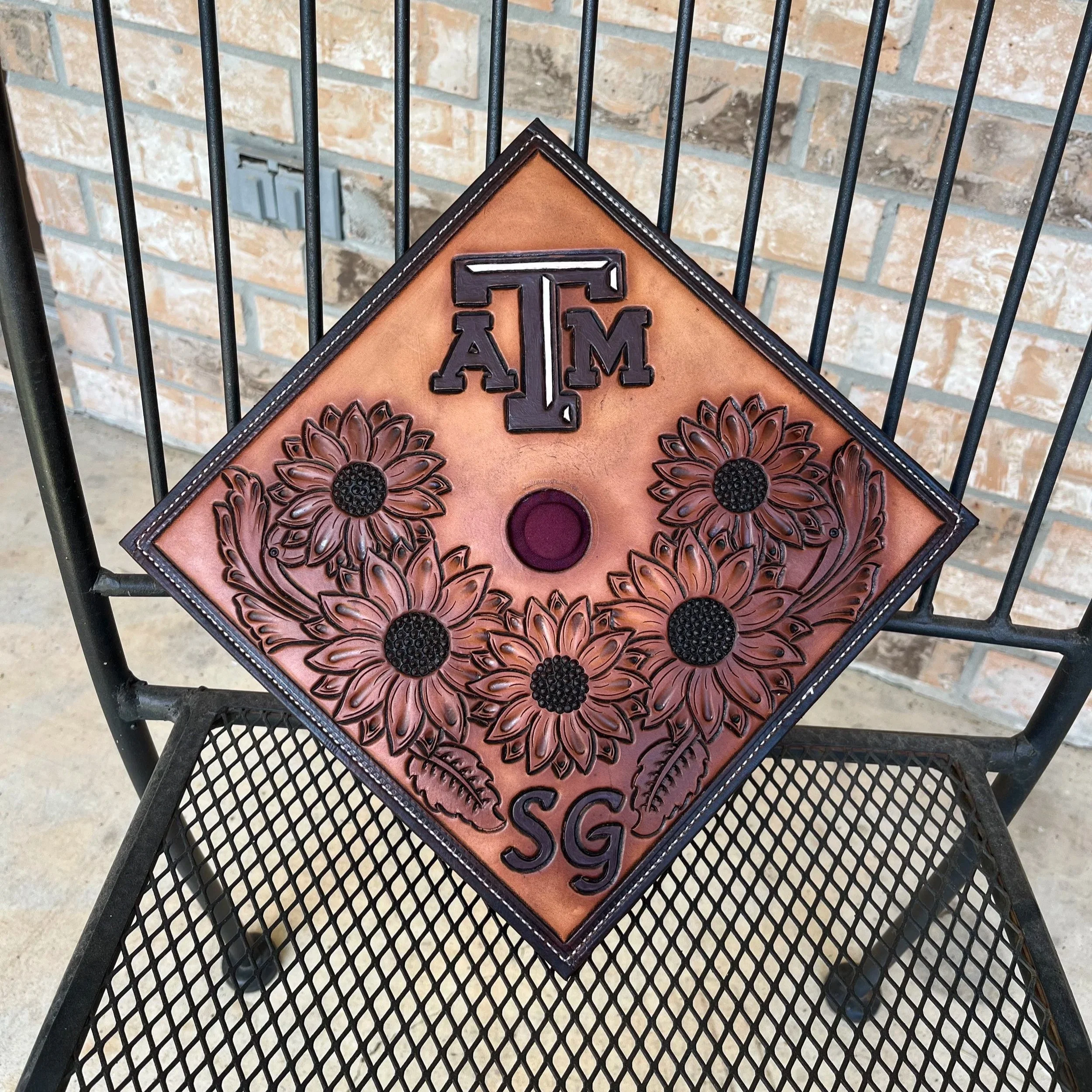Close-up of a decorative sign with the Texas A&M University logo and floral designs, placed on a black metal table with a brick wall in the background.