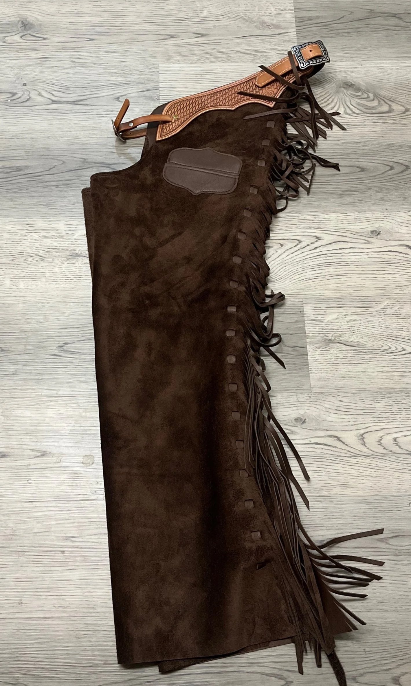 A pair of brown suede cowboy boots with fringes on the sides, laid on a light gray wooden floor.