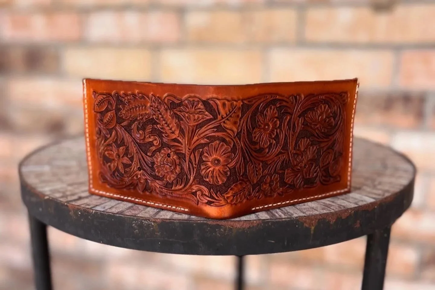 Leather wallet with intricate floral and leaf carvings, placed on a round wooden table with a brick wall background.