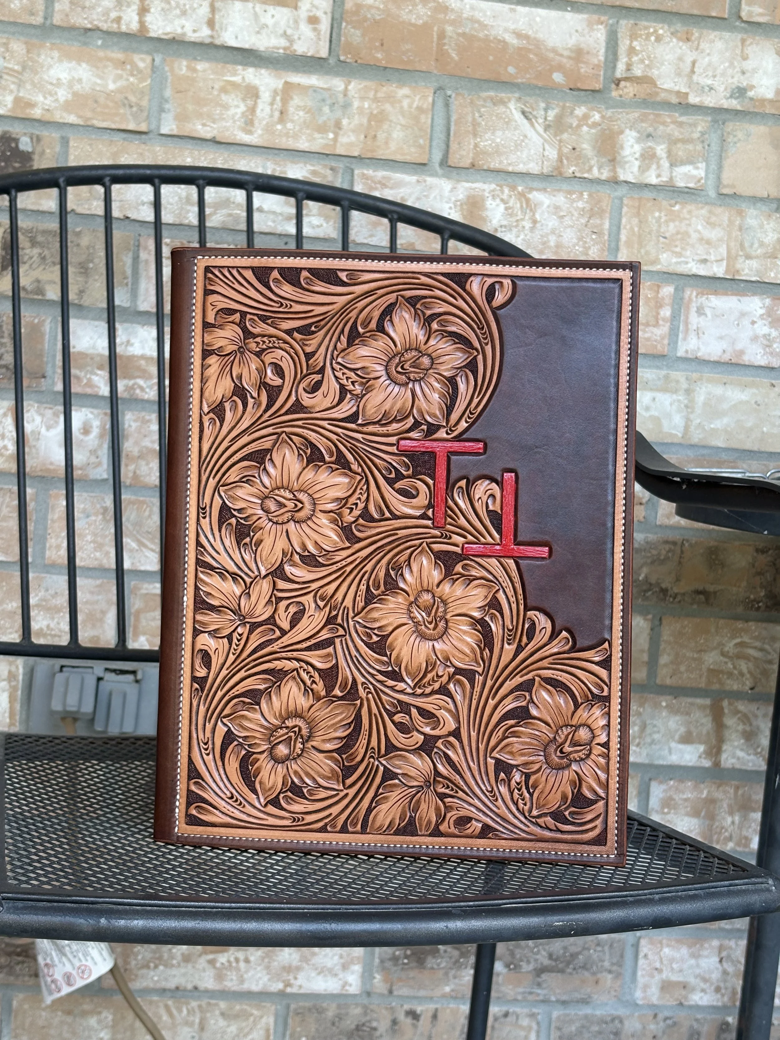 Leather-bound book with intricate floral carving and a red letter 'L' on the cover, resting on a metal outdoor table with a brick wall background.