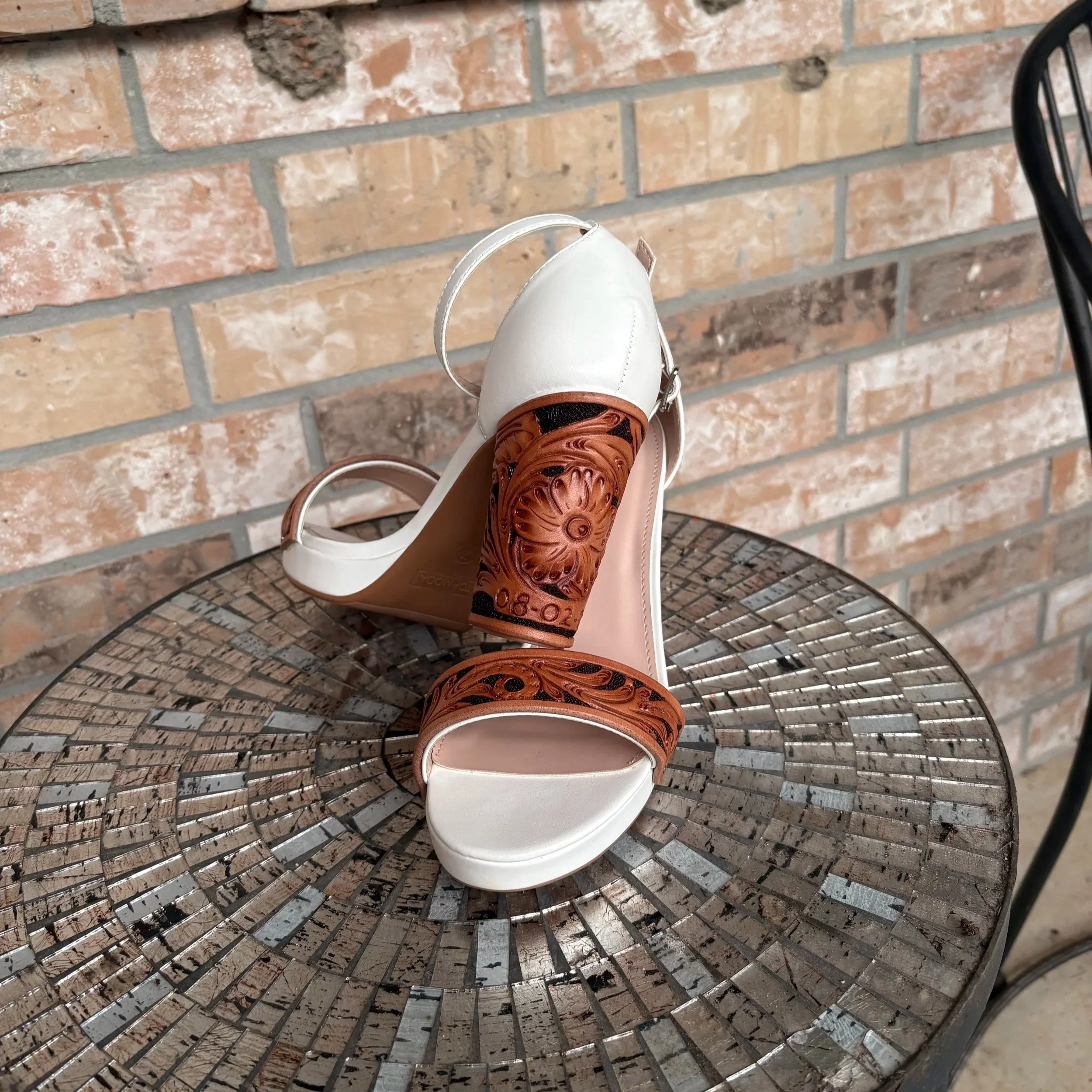 Photo of white high-heeled shoes with brown leather accents on a round metal table against a brick wall background.