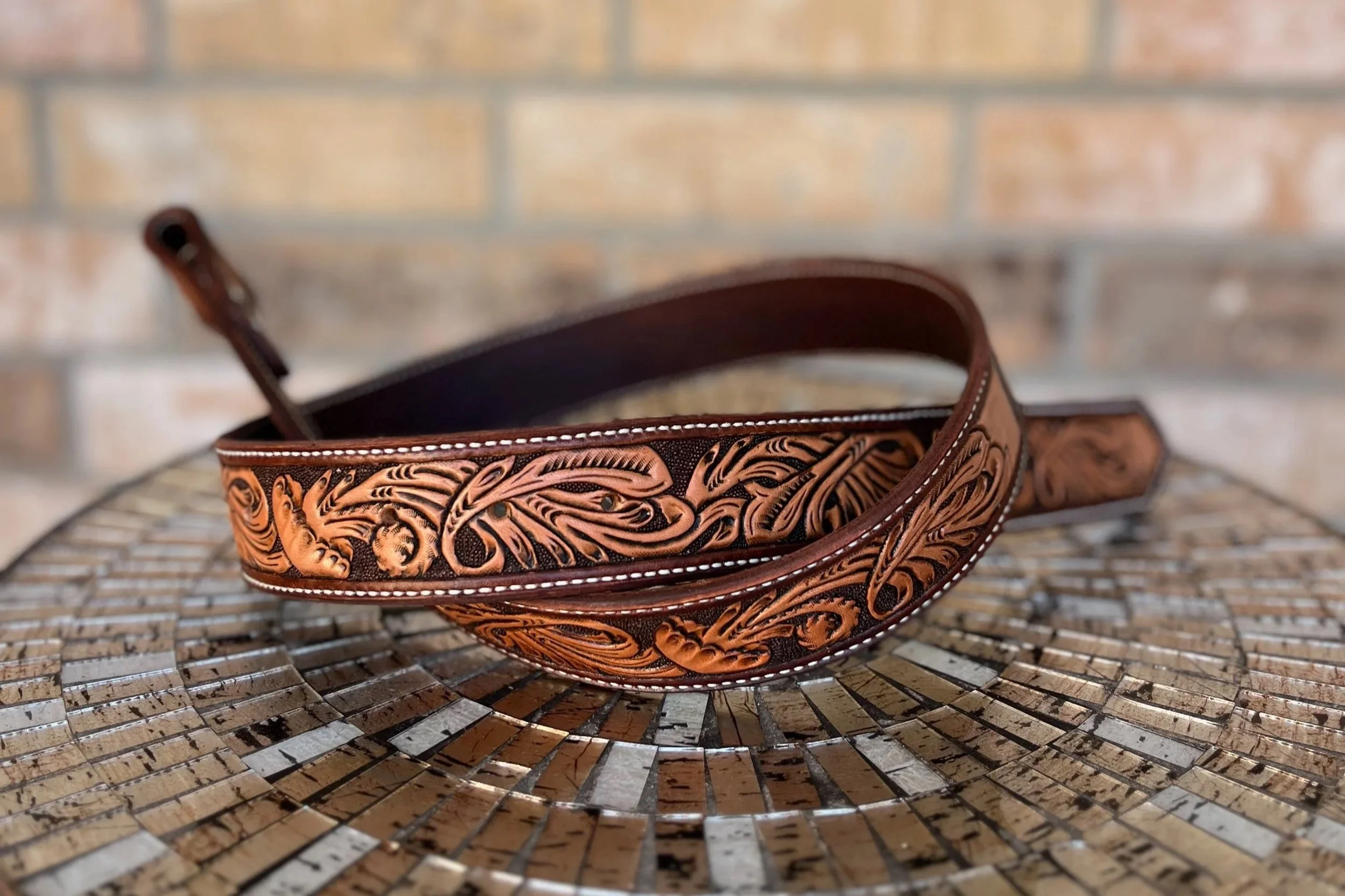 A brown leather belt with intricate carvings rests on a mosaic surface with a brick wall background.