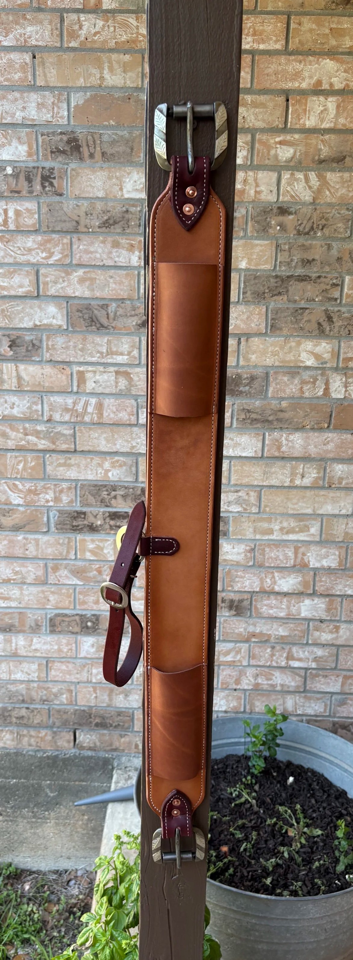 Leather rifle sling hanging on a wooden post against a brick wall, with a garden pot and plants at the base.