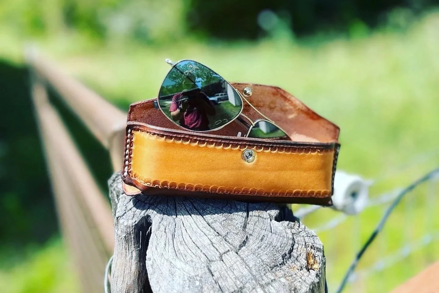 A pair of sunglasses resting in an orange leather case on a weathered wooden post outdoors during daytime.