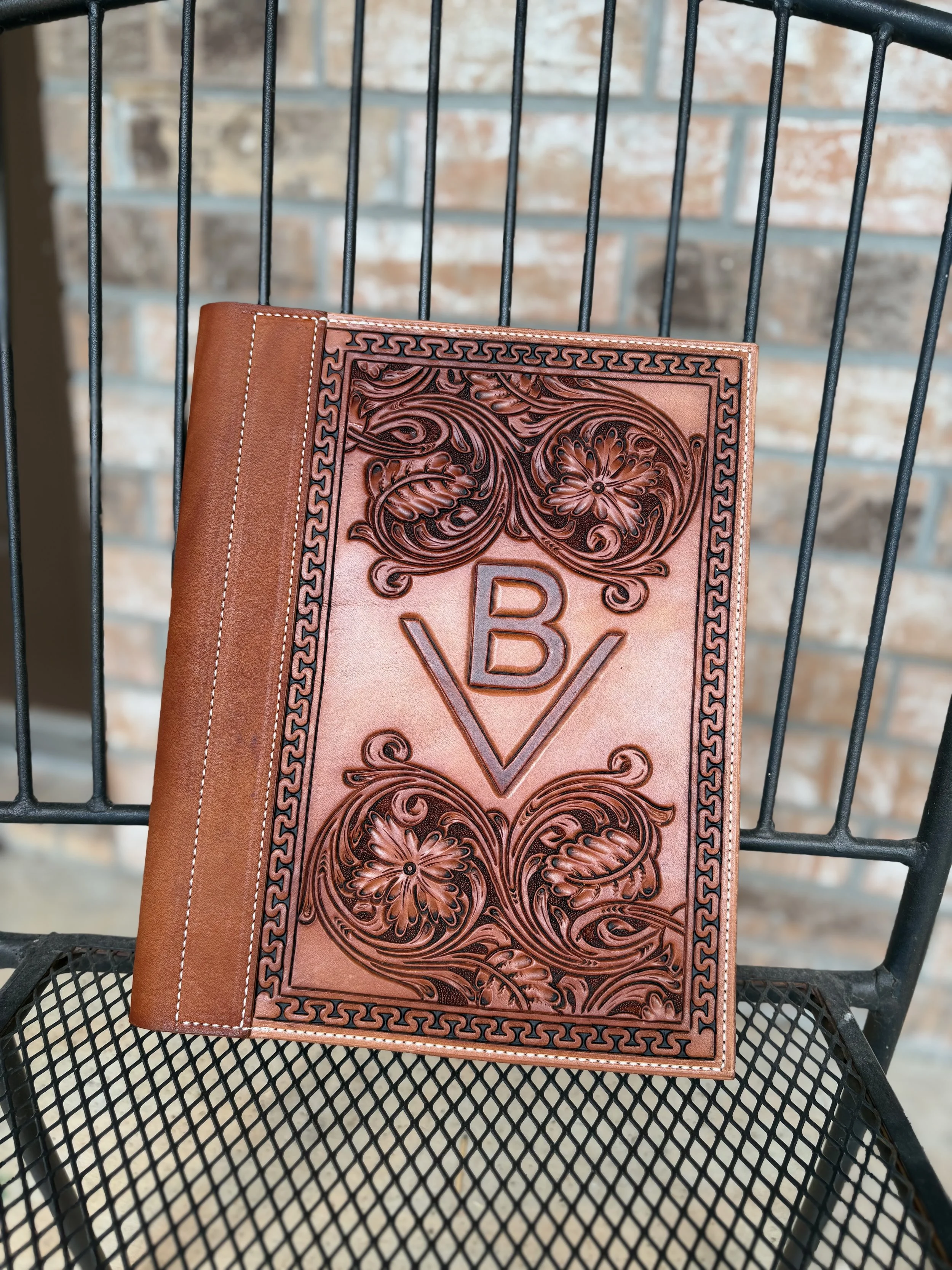 A leather-bound book or journal with intricate floral and geometric embossing on the cover, resting on a black metal wire outdoor table with a brick wall in the background.