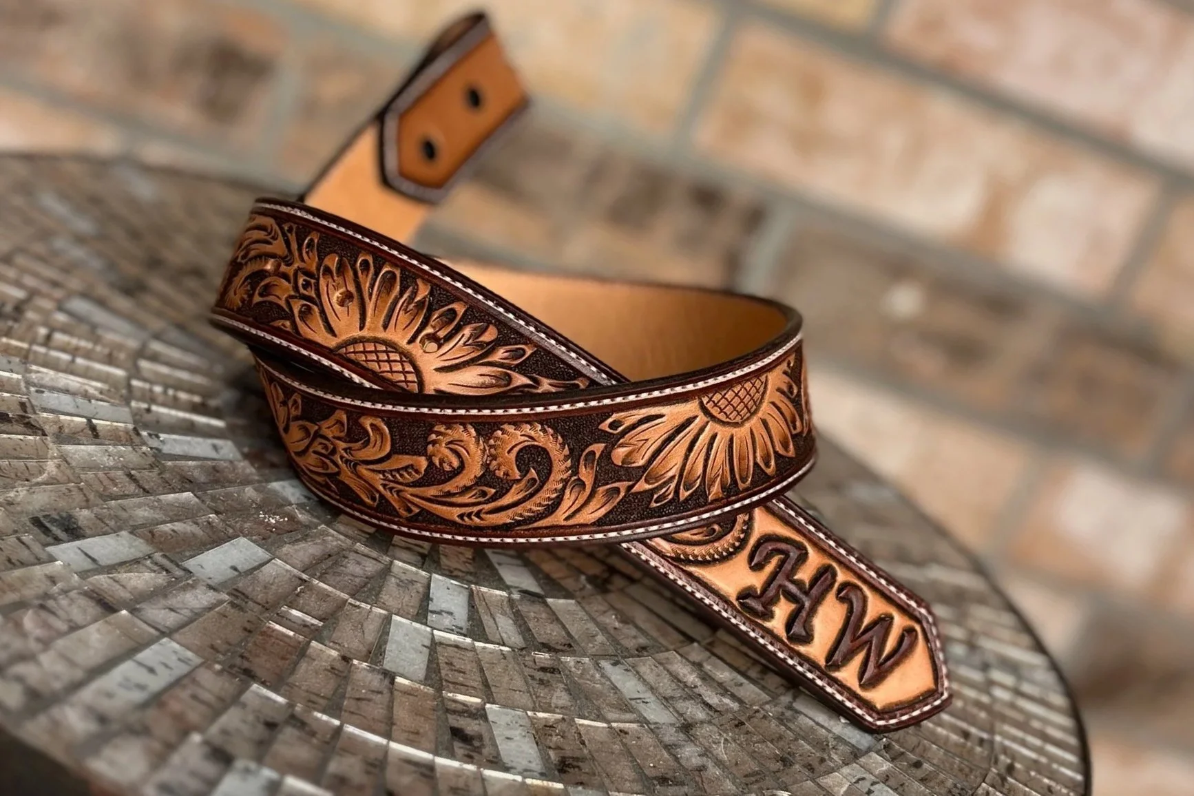 A leather belt with intricate floral and swirl carved patterns, resting on a woven surface with a mismatched brick wall background.