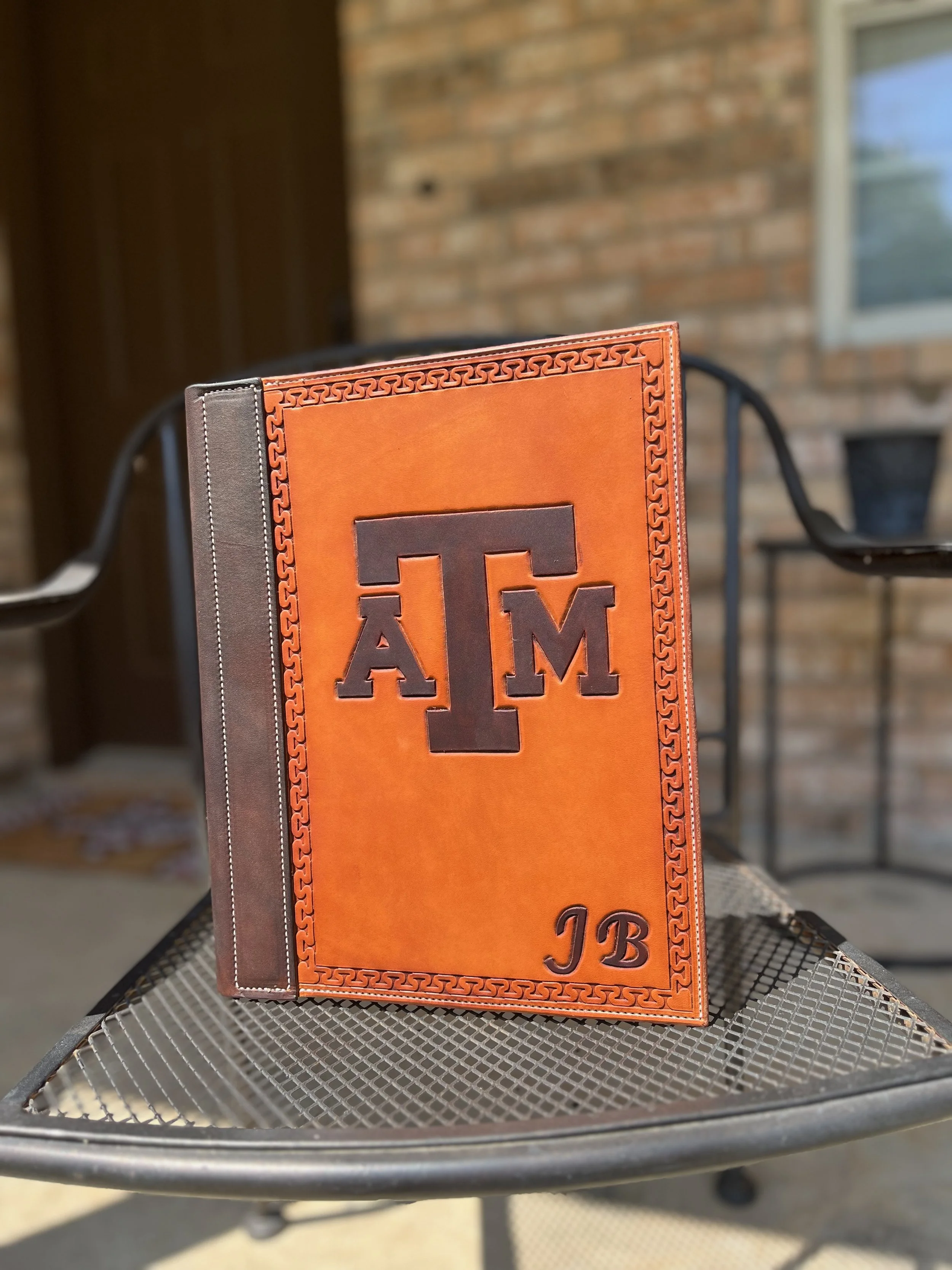 A Texas A&M University 12th Man hardcover book with an orange cover and black lettering, placed on a metal outdoor table with a brick wall and window in the background.
