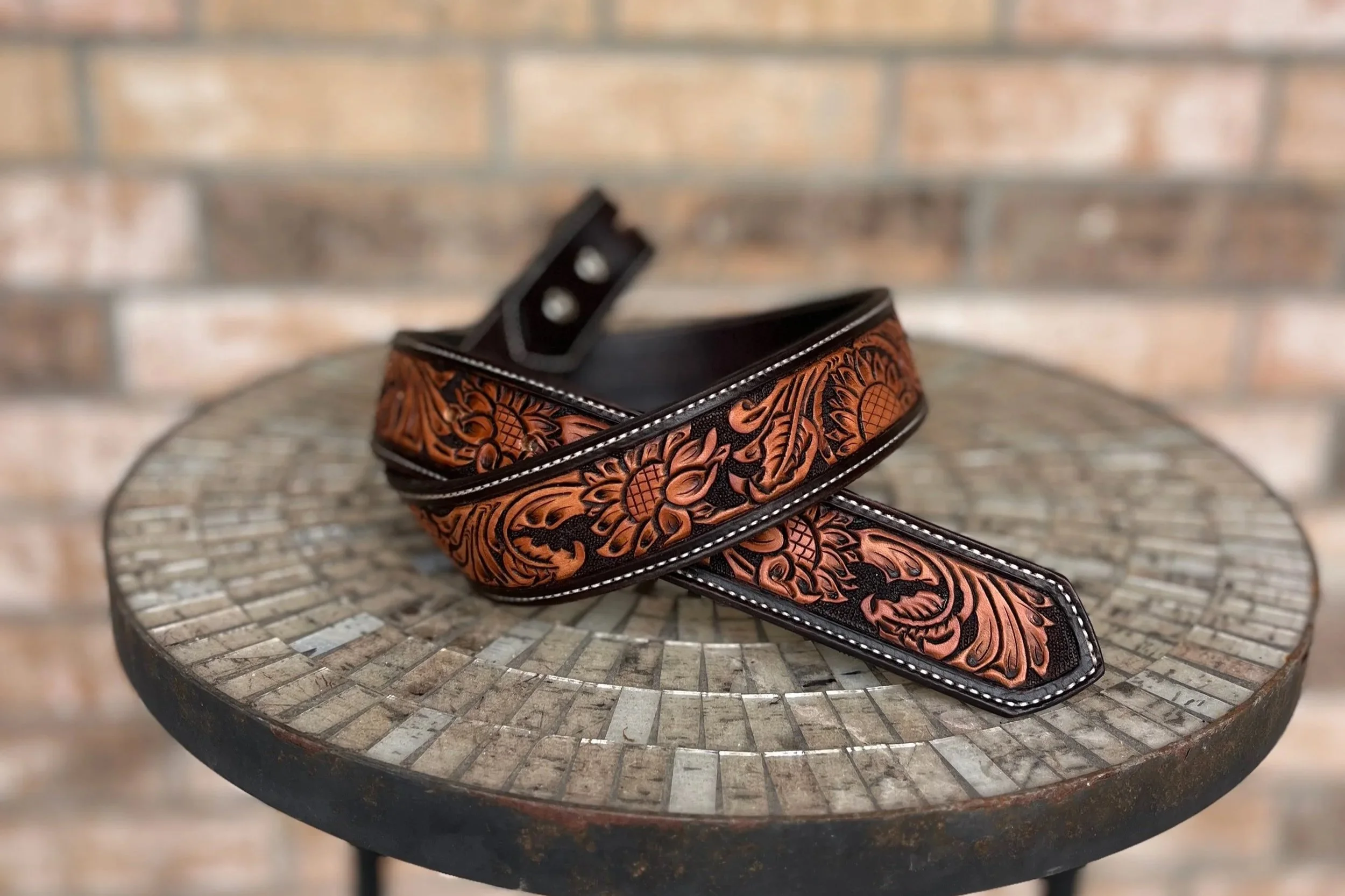 Leather belt with intricate floral and geometric patterns on a black background, placed on a round metal table against a blurred brick wall.
