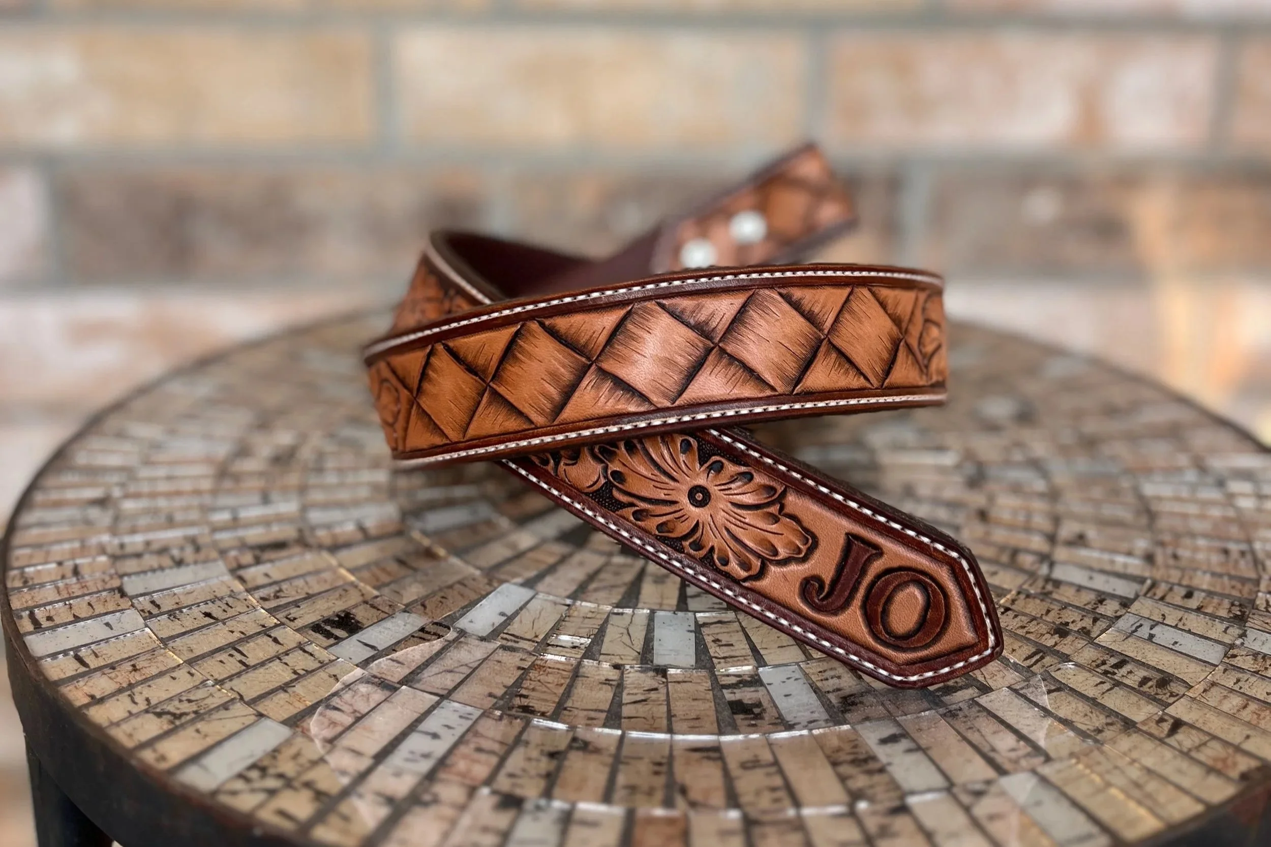 A brown leather belt with decorative tooling and silver buckle details, resting on a round mosaic table with a brick wall in the background.