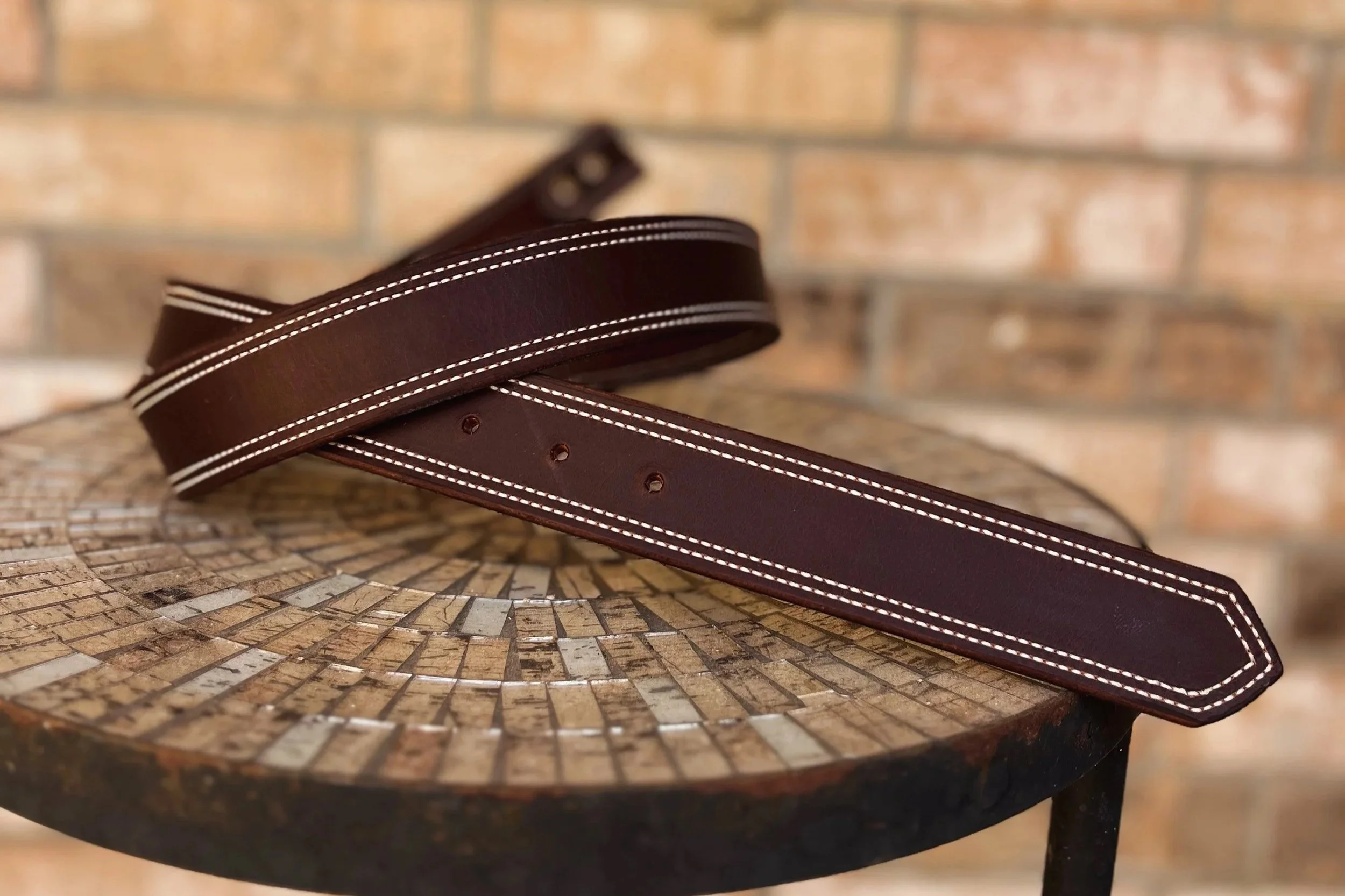 A close-up of a brown leather belt with white stitching, draped over a mosaic-tiled round table with a brick wall in the background.