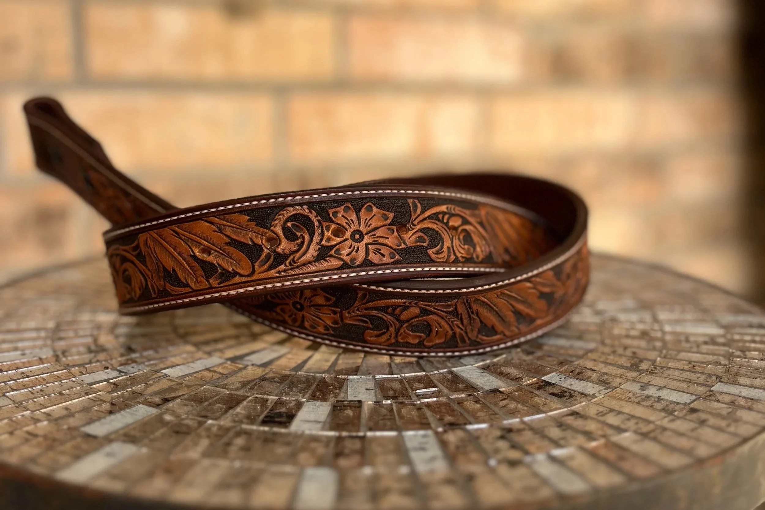 A leather belt with intricate floral carving lies coiled on a mosaic-style surface, with a blurred brick wall background.