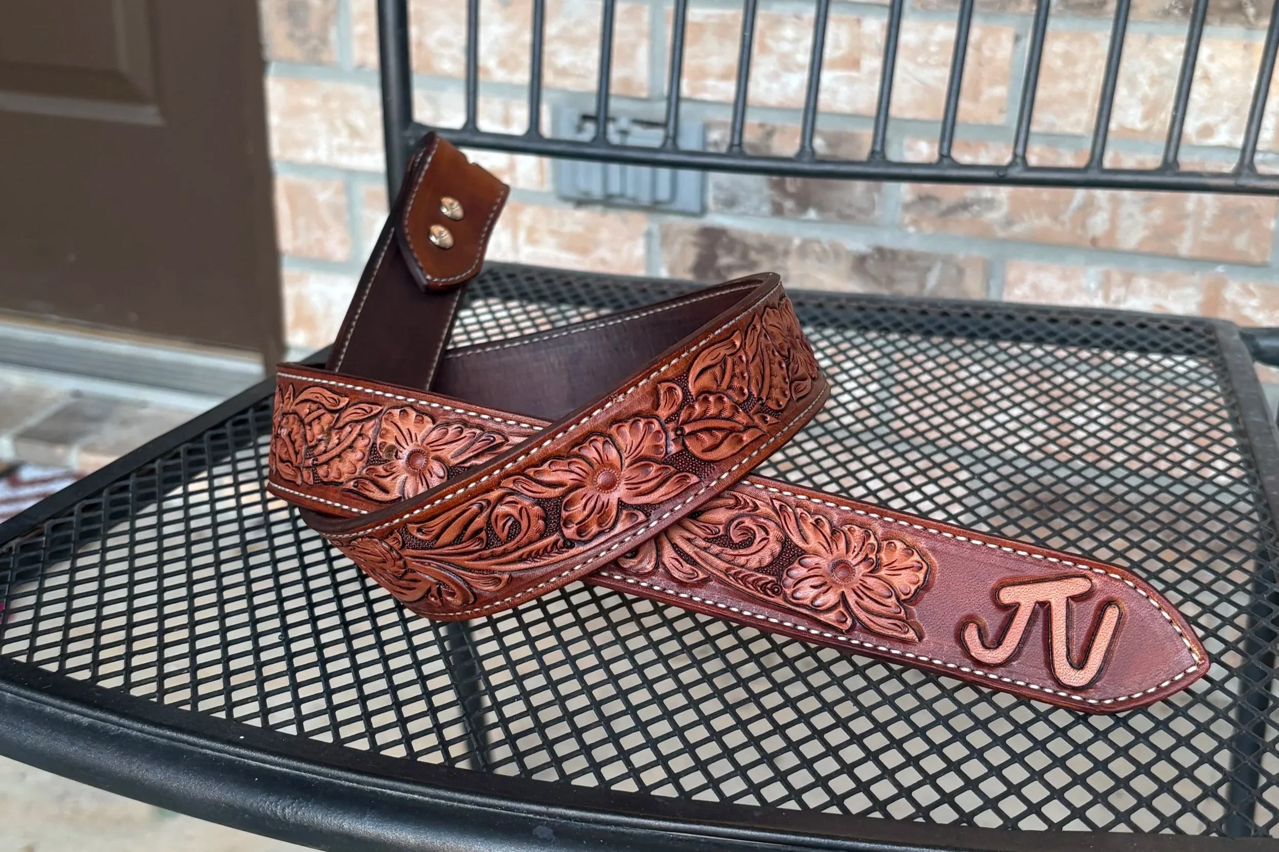 A brown leather belt with floral and swirl pattern carved into it, resting on a black metal mesh outdoor table, with brick wall and metal railing in the background.