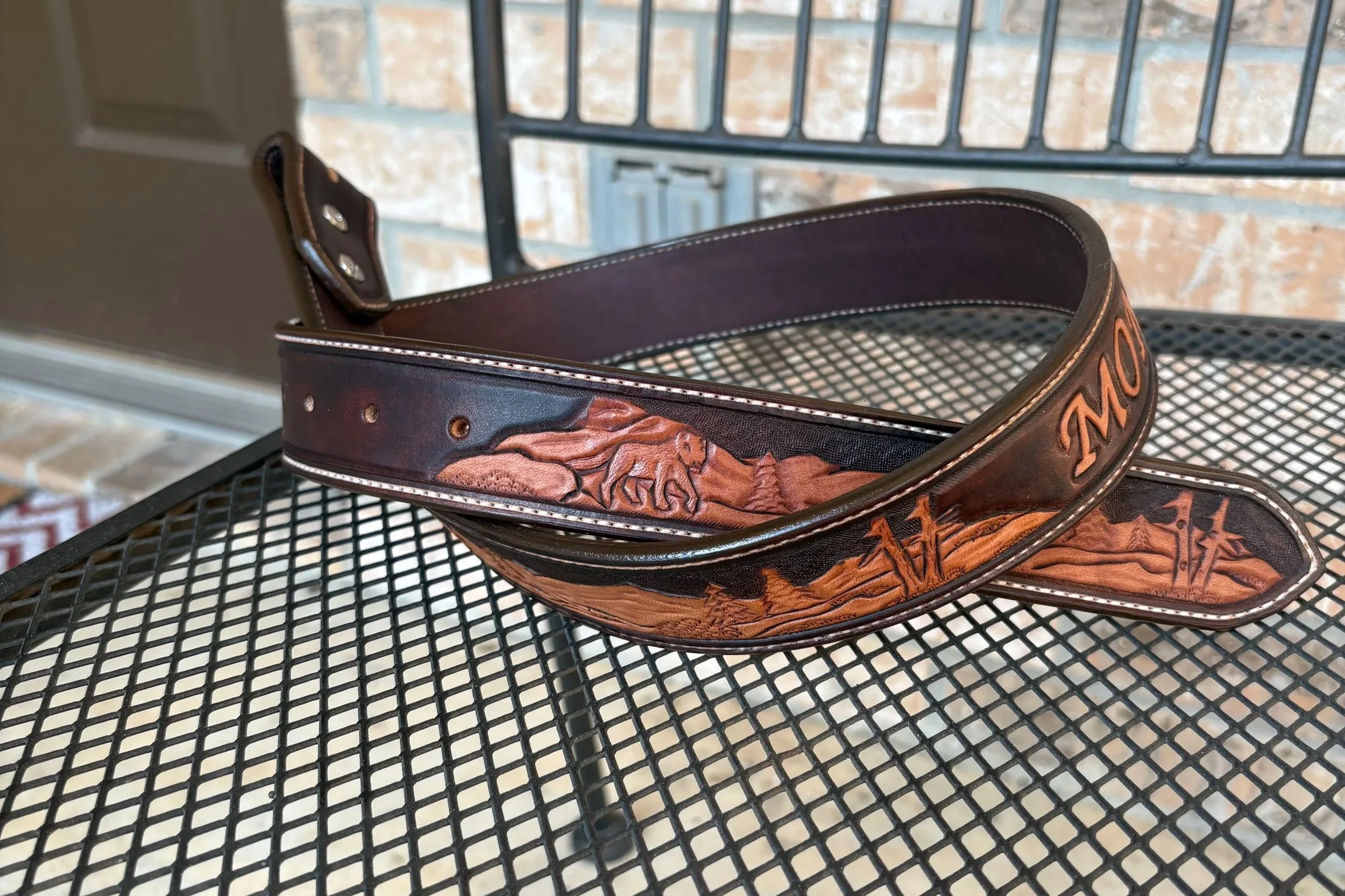 Leather golf belt with embossed mountain and animal designs, resting on a metal mesh table with a brick wall and metal gate in the background.