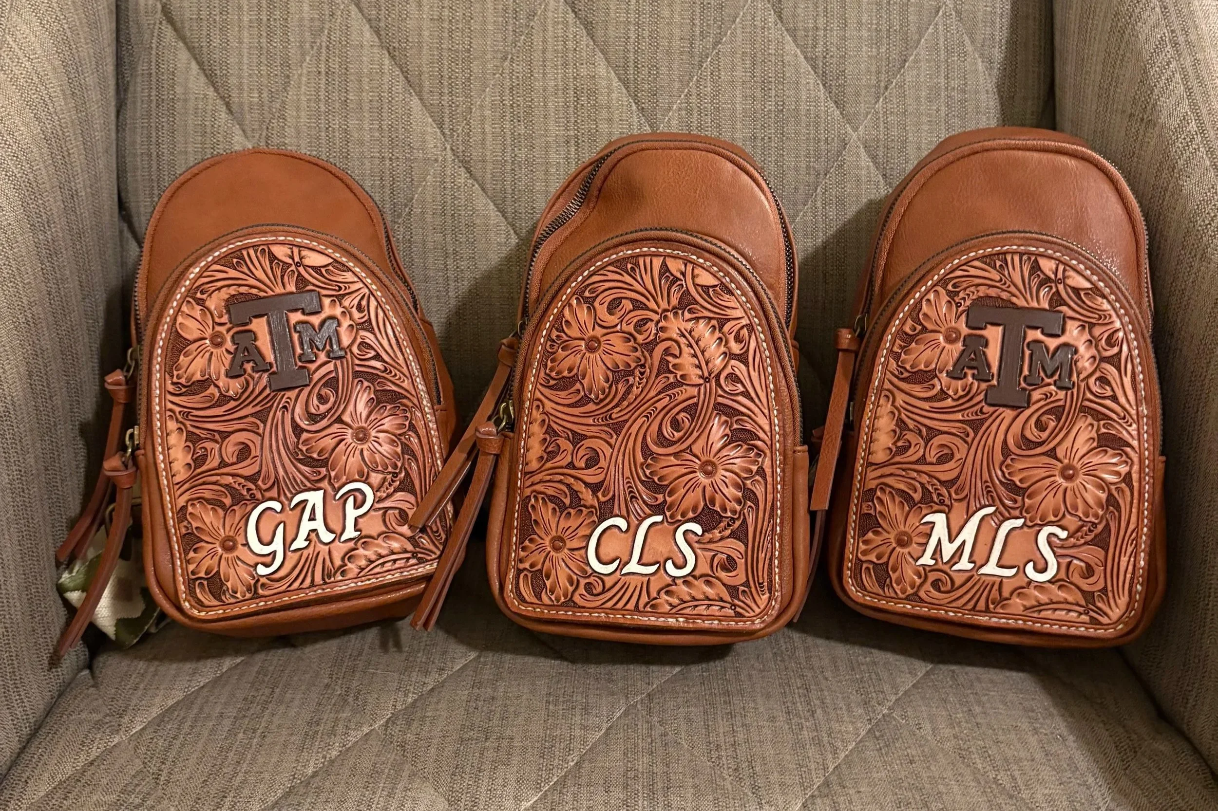Three embroidered backpacks with floral patterns and university initials, placed on a beige textured chair.