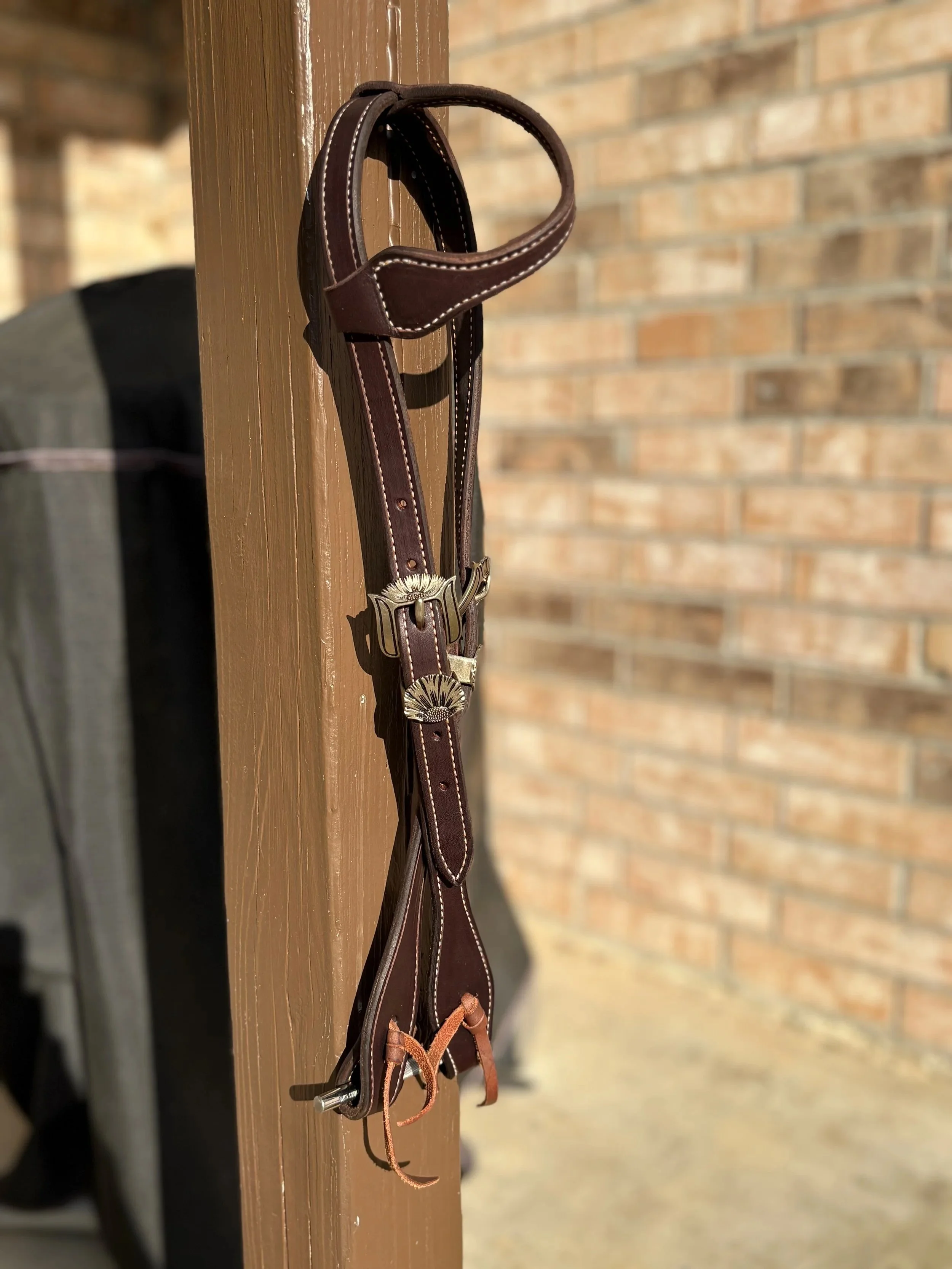 A leather horse bridle hangs on a corner of a wooden wall with a brick wall in the background.