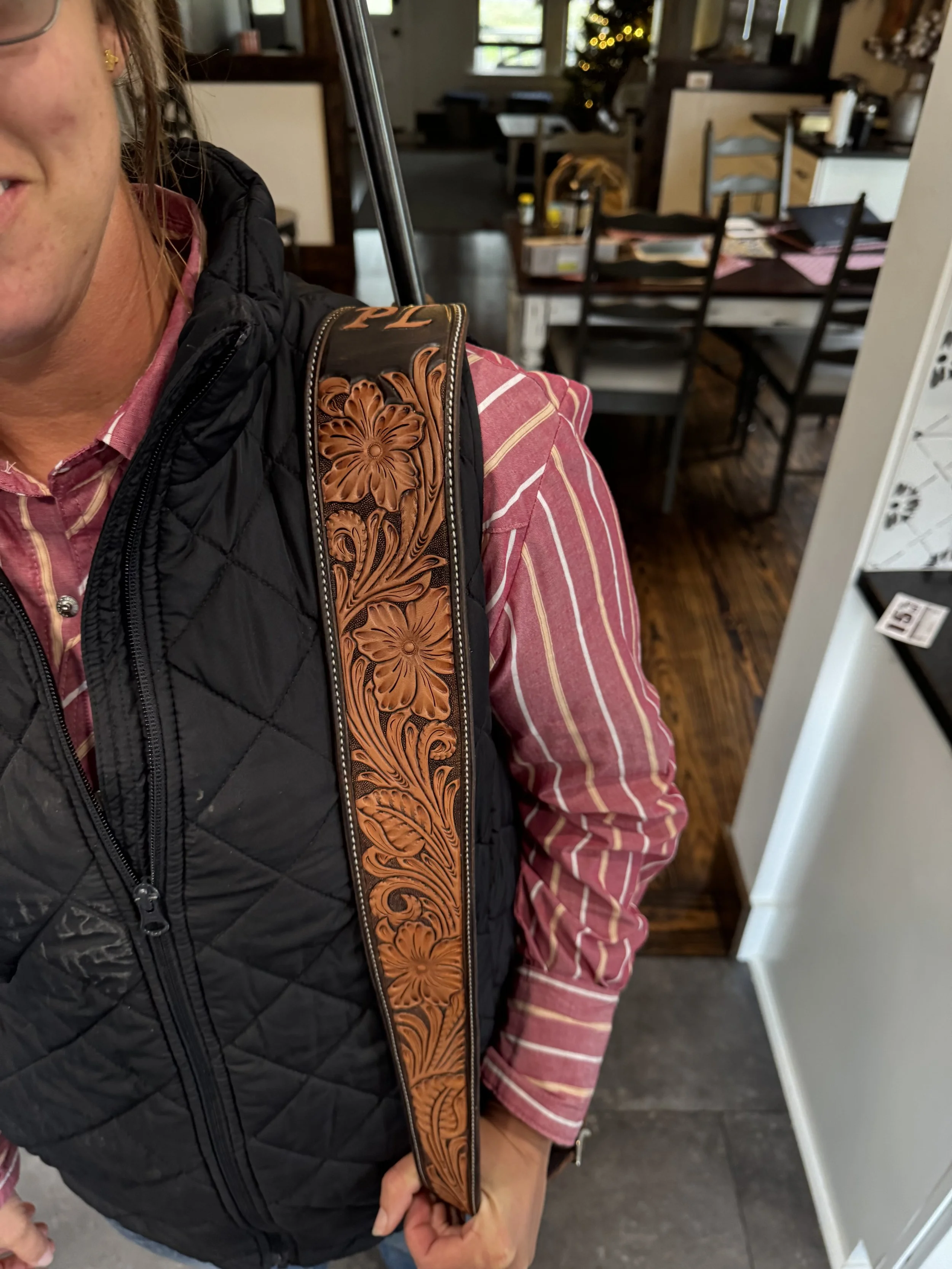 Part of a person’s face and arm, wearing a pink and white striped shirt, black quilted vest, and carrying a leather strap with floral embossing, in a room with dining table, chairs, and a decorated Christmas tree.