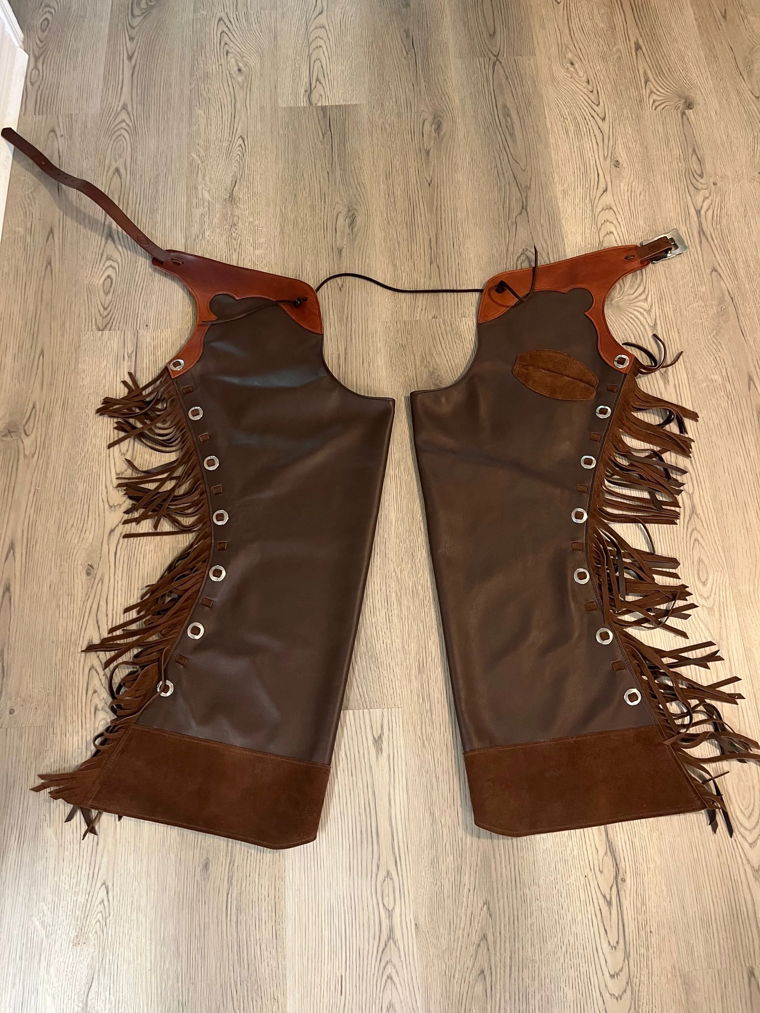 A pair of brown leather fringe chaps with metal eyelets and a leather patch on each leg, laid flat on a wooden floor.