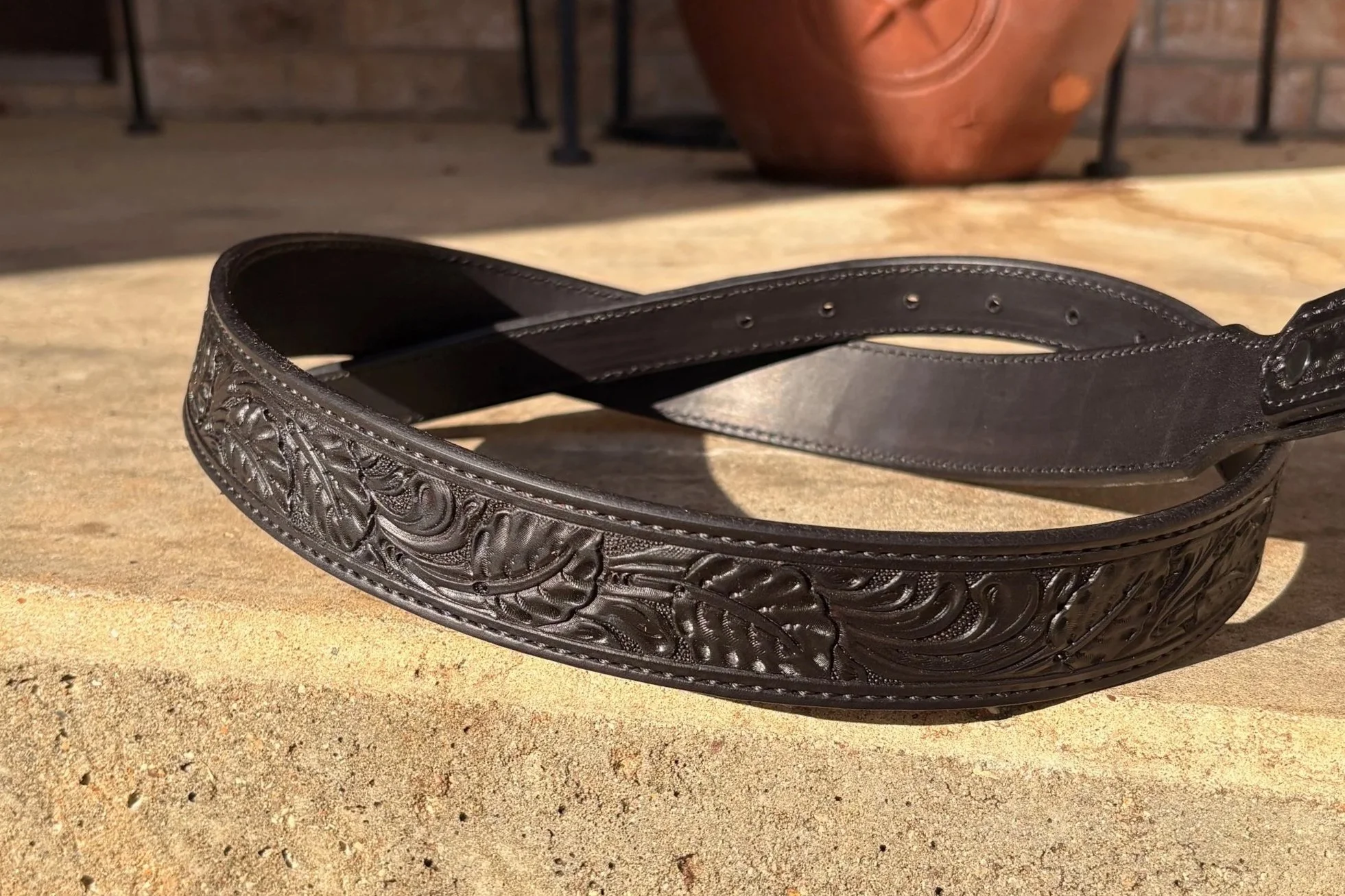 A black leather belt with intricate floral and swirl embossing lying on a stone surface, with a large terracotta pot and part of a metal railing in the background.